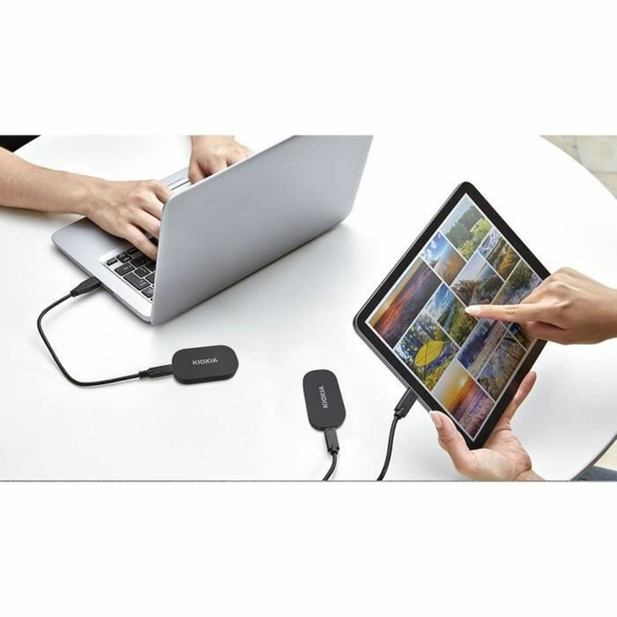 Ein Laptop und ein Tablet sind über schwarze Kabel mit KIOXIA-Geräten verbunden. Eine Person benutzt jedes Gerät, beide auf einem weißen Tisch.