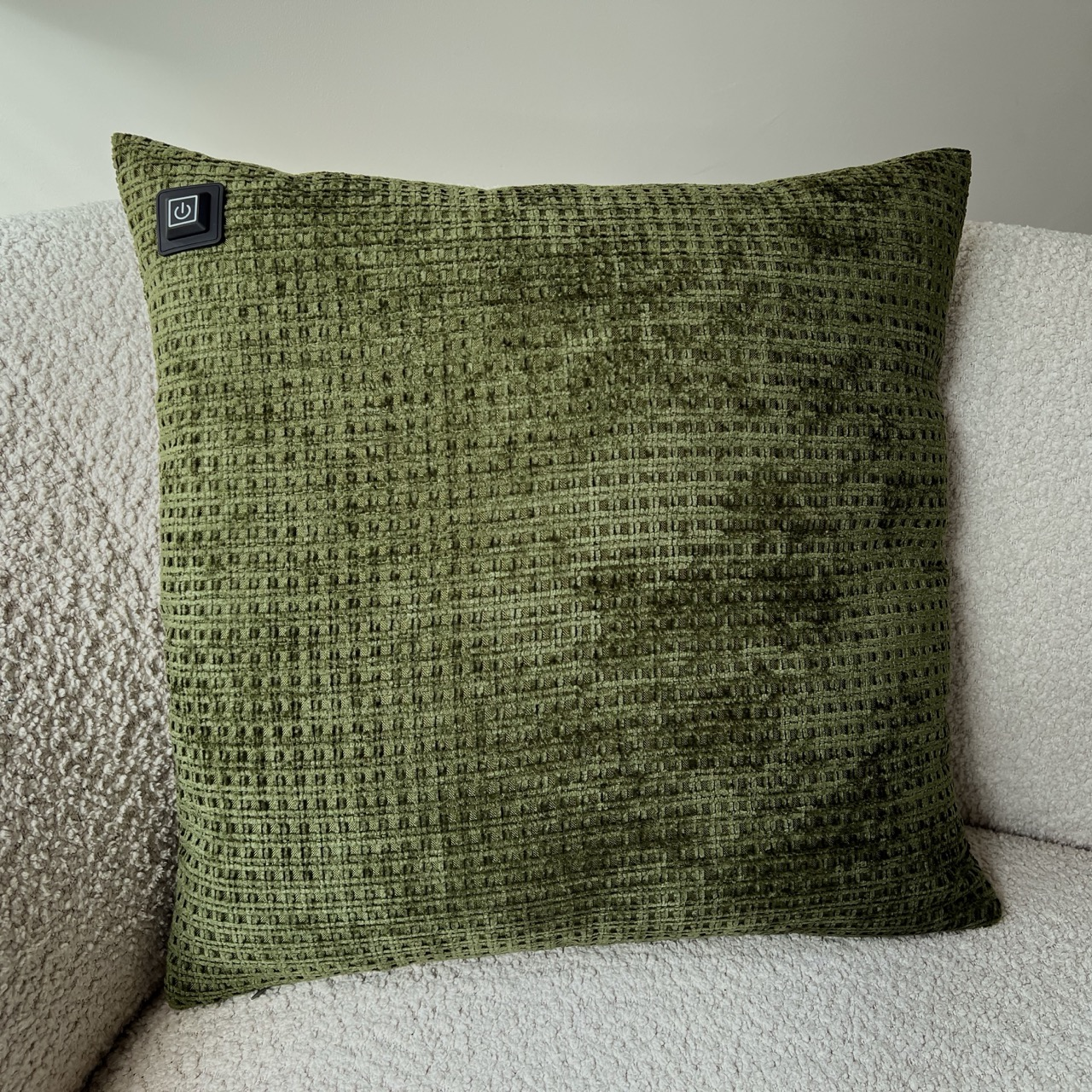Ein grünes, strukturiertes Kissen mit einem Power-Knopf liegt auf einem weißen, flauschigen Sofa. Das Kissen hat ein quadratisches Muster.