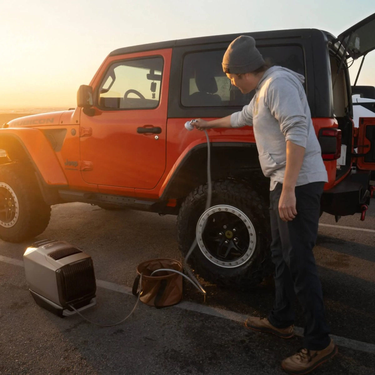 Eine Person wäscht einen orangenen Jeep mit einem Schlauch. Ein tragbarer Wasserkocher und ein Eimer stehen auf dem Boden. Der Hintergrund ist ein Sonnenuntergang.