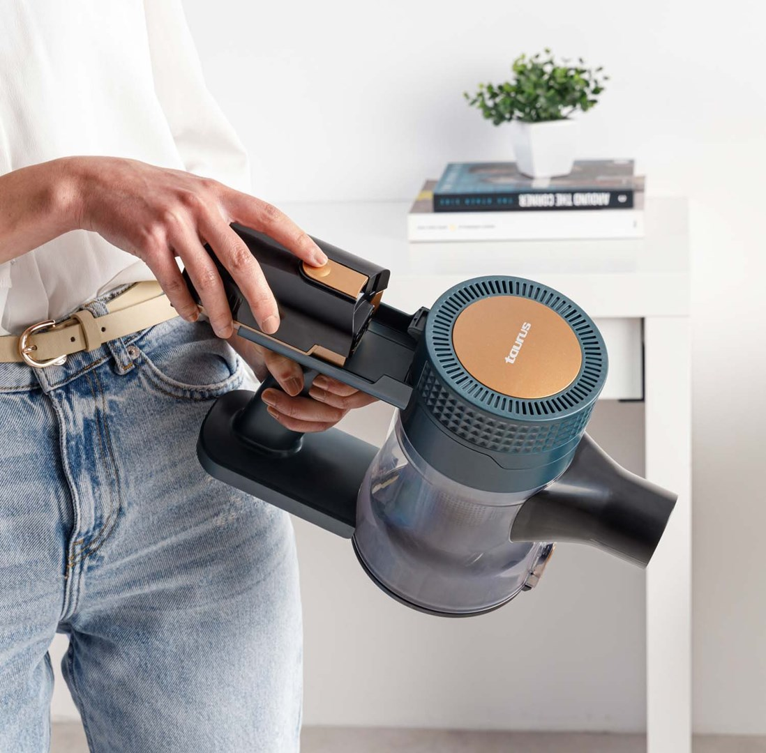 Osoba trzyma bezprzewodowy odkurzacz. Odkurzacz jest czarny i morski, ze złotym akcentem. Biały stół i roślina w tle.