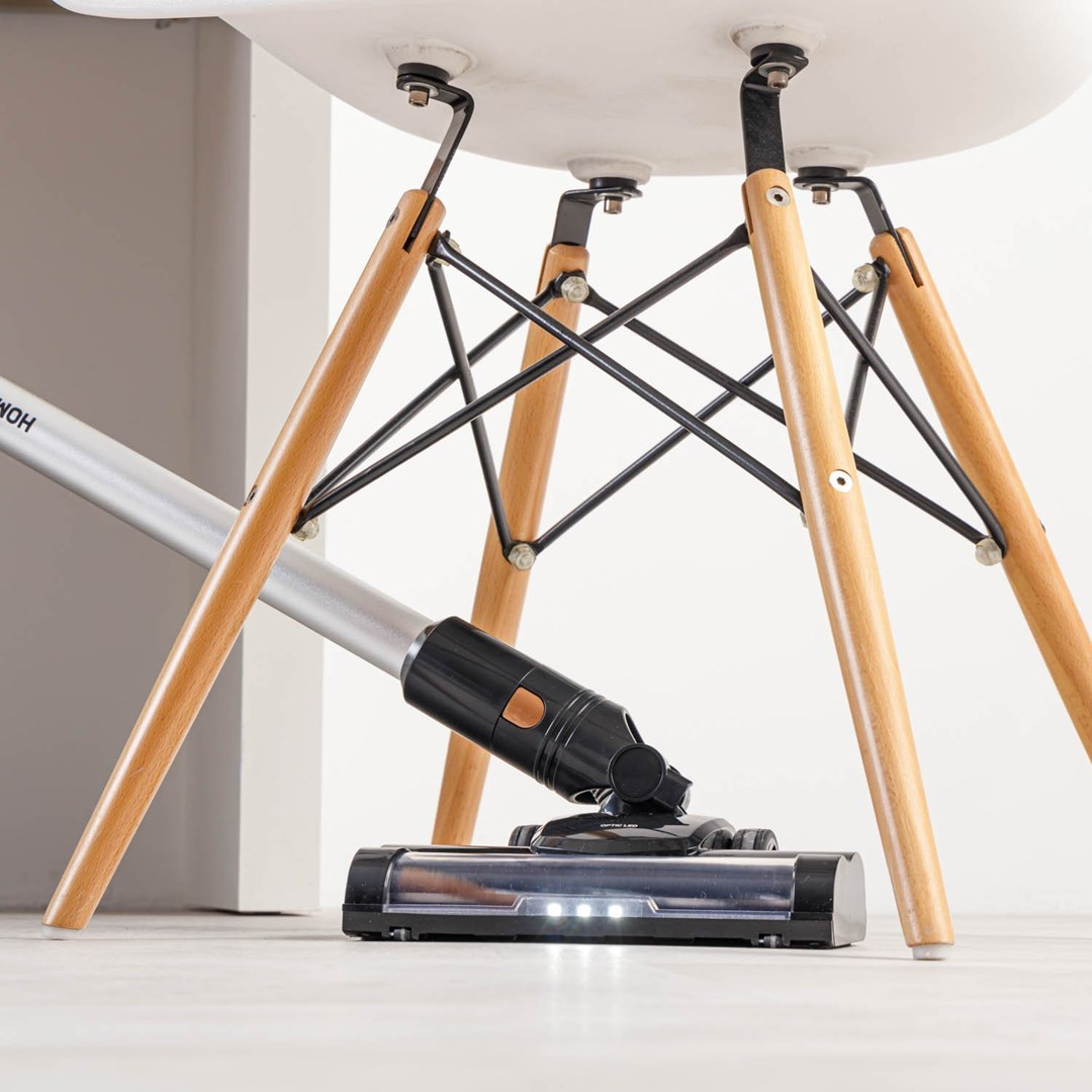 Odkurzacz czyści pod białym krzesłem. Krzesło ma drewniane nogi i czarne metalowe podpory.