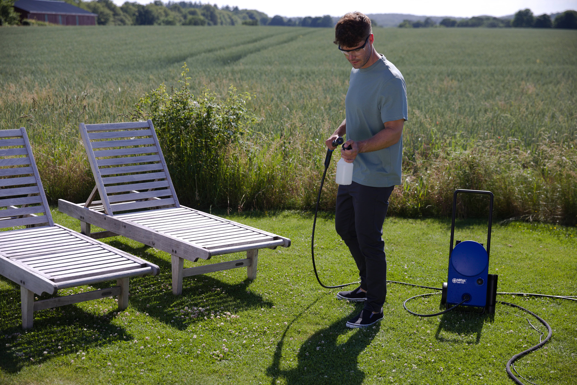Mężczyzna z myjką ciśnieniową obok dwóch drewnianych leżaków na trawie. Pole i budynek w tle.