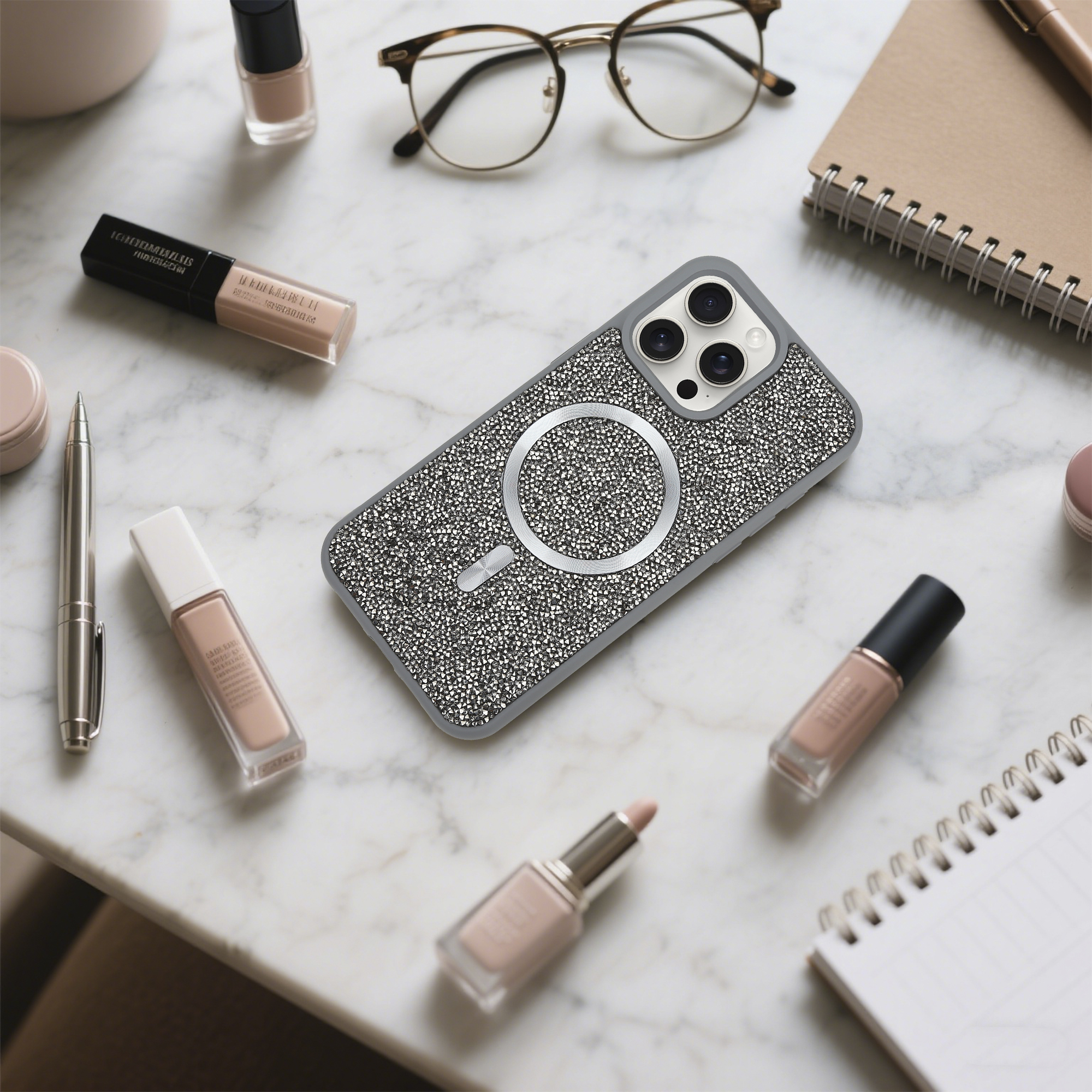 Ein Telefon in einer verzierten Hülle, eine Brille, Make-up, Nagellack, ein Stift und ein Notizbuch liegen auf einer Marmoroberfläche.