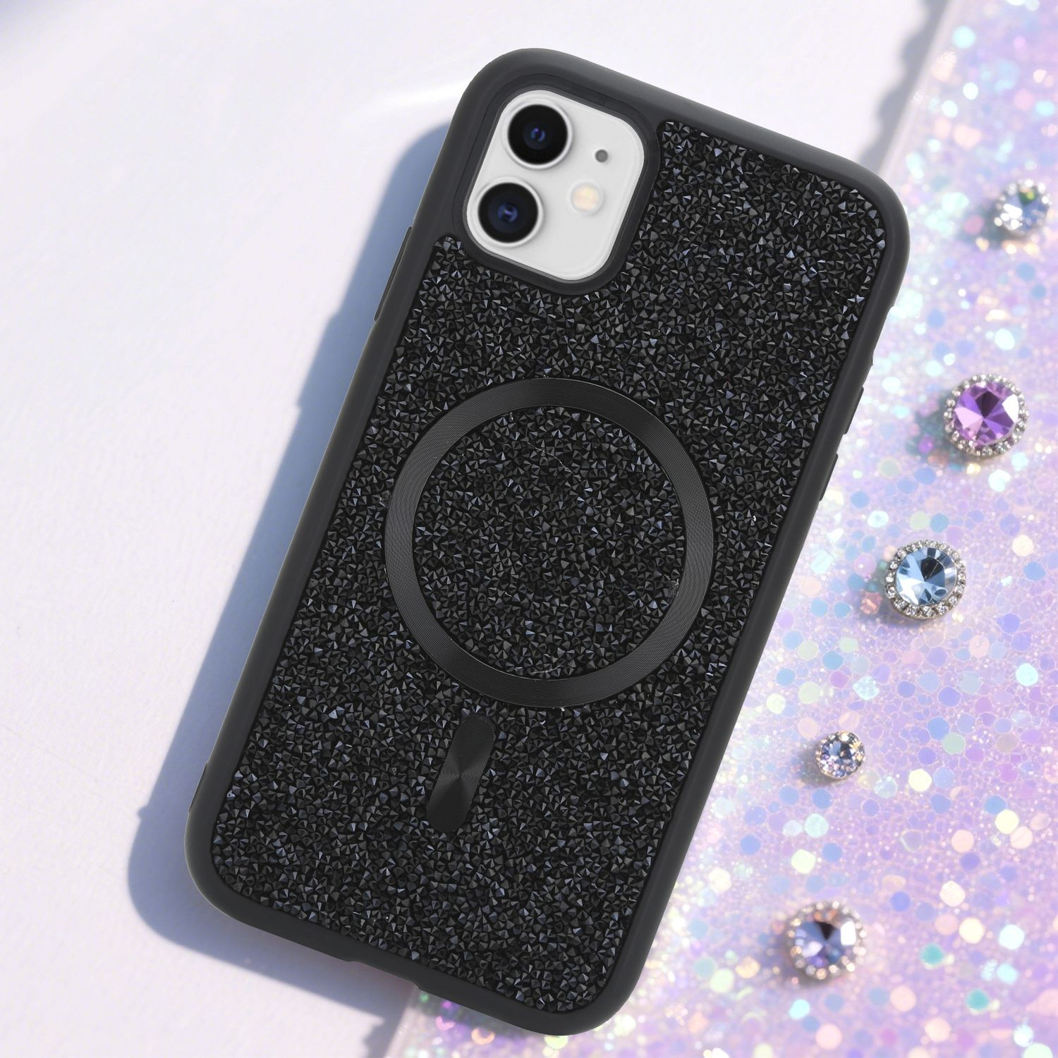 Eine schwarze Handyhülle mit kleinen schwarzen Kristallen und einem Magnetring liegt auf einer glitzernden Oberfläche mit Edelsteinen.
