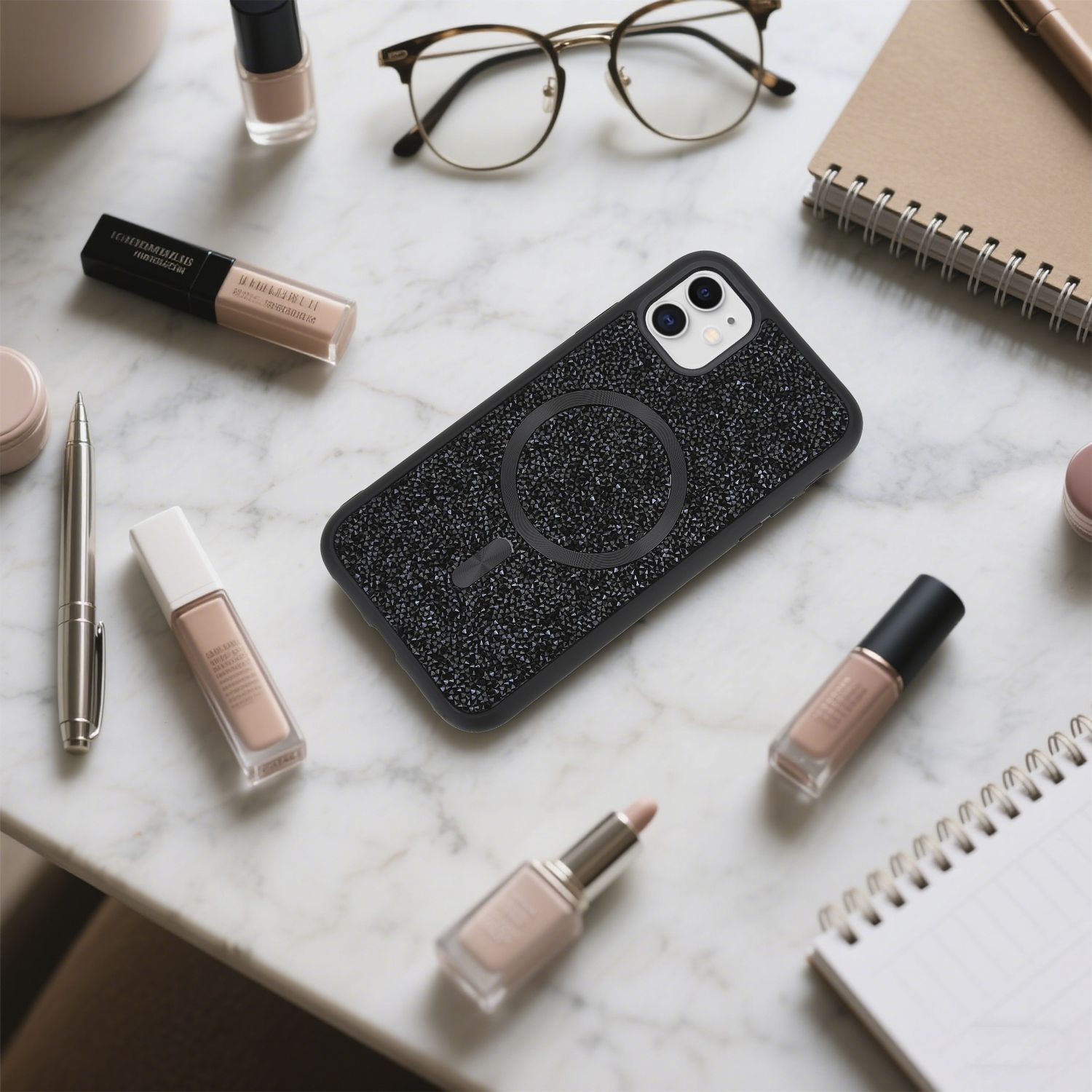 Auf einer Marmoroberfläche liegen eine Handyhülle, Make-up, eine Brille und ein Notizbuch. Die Handyhülle ist schwarz und glitzernd. Make-up in verschiedenen Farbtönen.