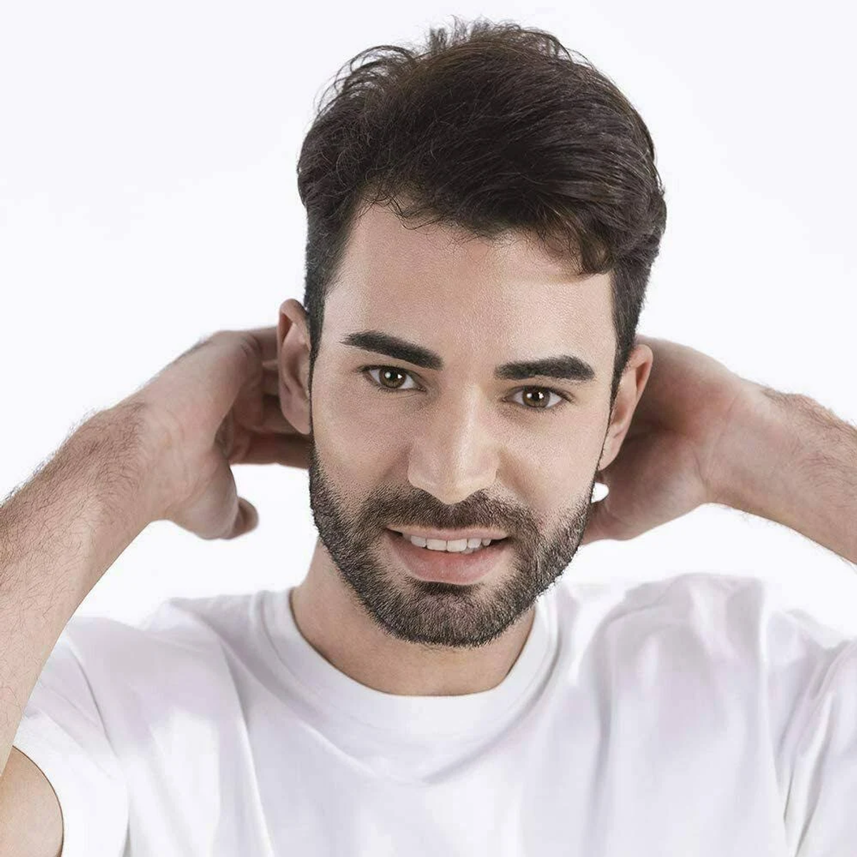 Un hombre con cabello oscuro y barba sonríe, manos detrás de la cabeza. Lleva una camiseta blanca. Fondo blanco.