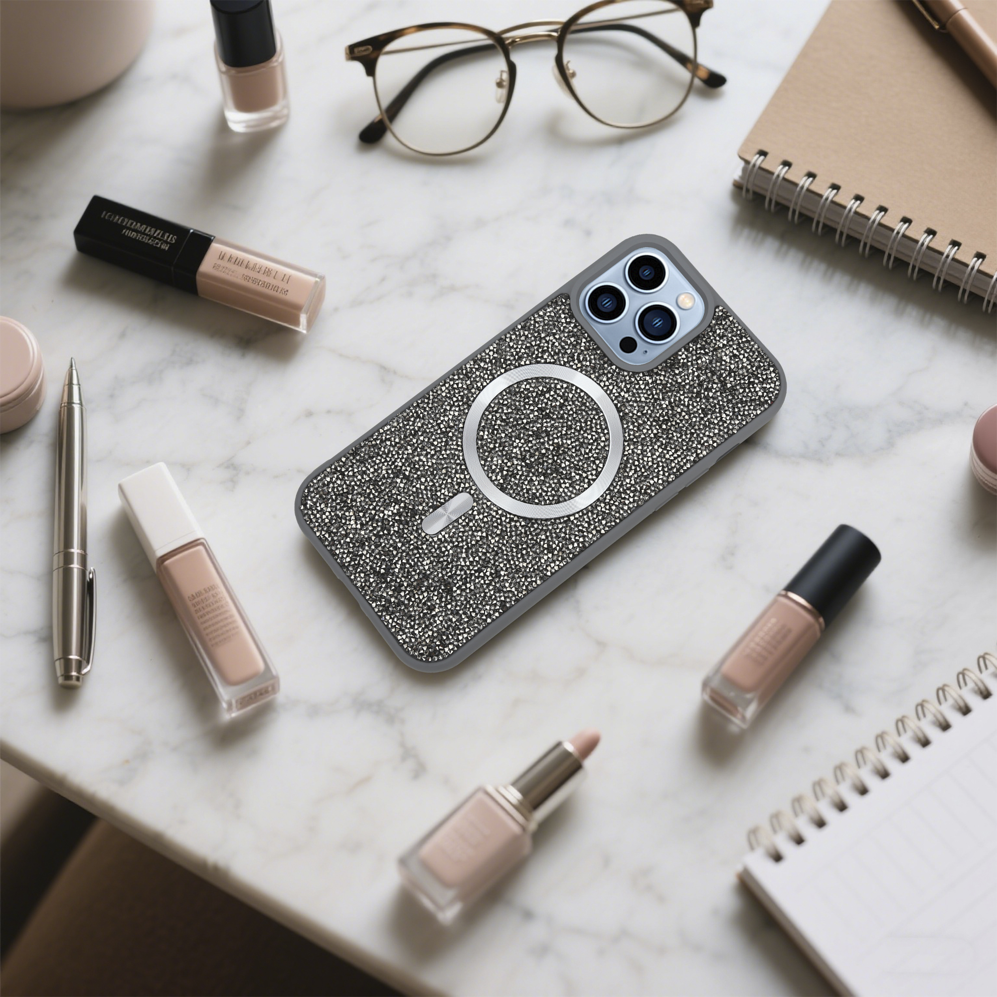 Ein Telefon in einem verzierten Gehäuse, eine Brille, Make-up und ein Notizbuch auf einer Marmoroberfläche.