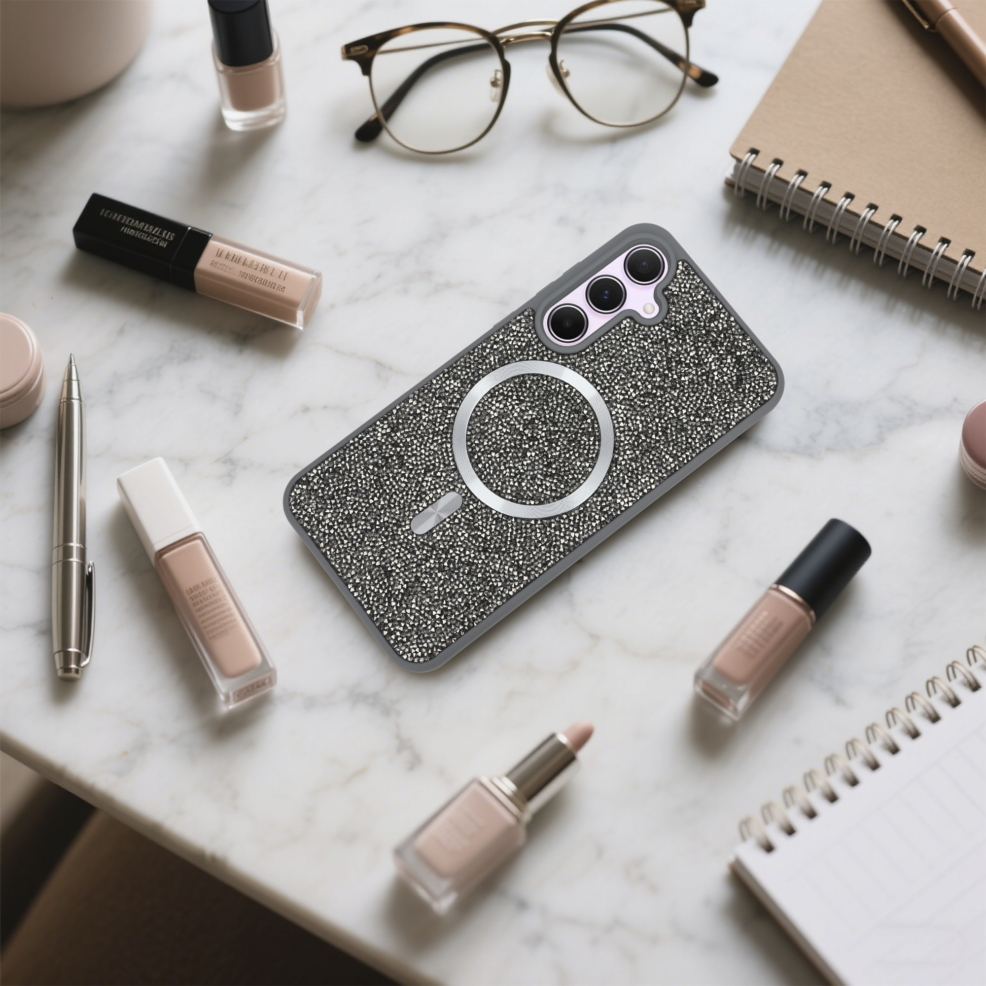 Auf einer Marmoroberfläche befinden sich eine Handyhülle, Make-up, eine Brille und ein Notizbuch. Die Handyhülle ist mit Strass besetzt.