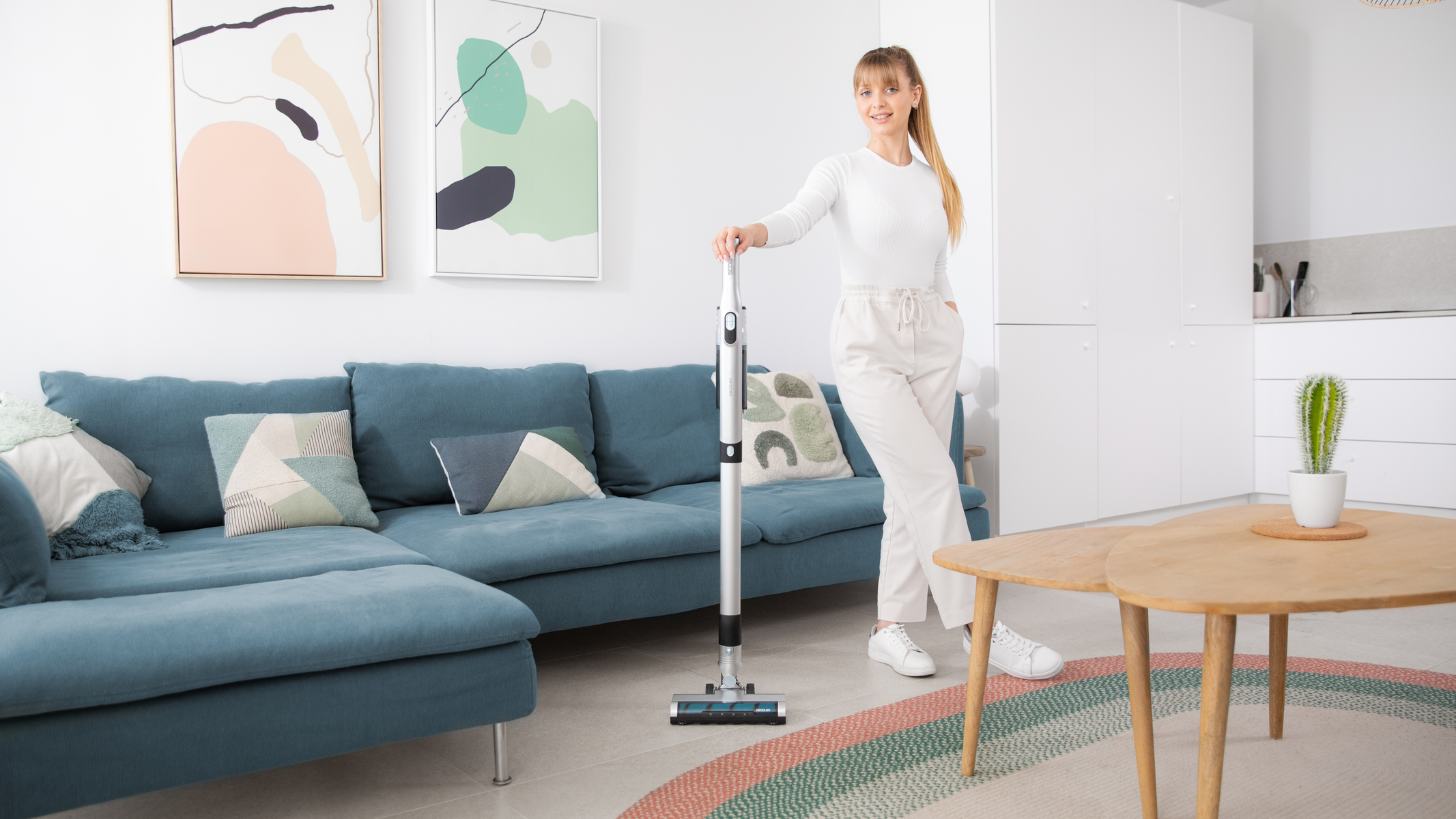 Mujer de blanco con aspiradora en salón moderno. Sofá azul, arte, mesa de madera y cactus.