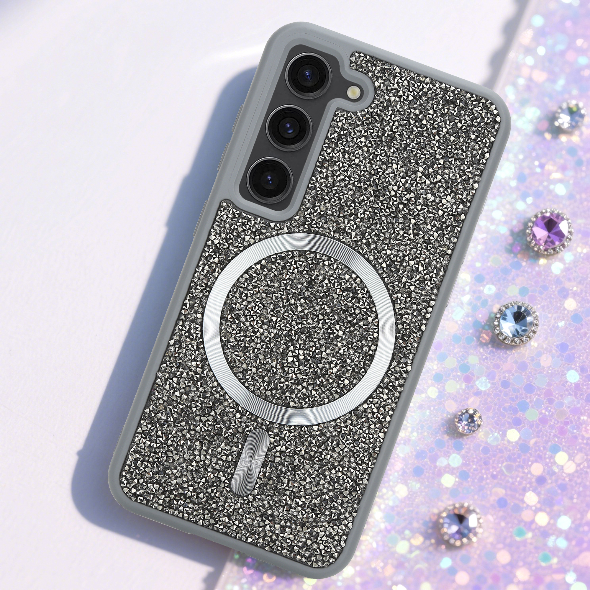 Ein verziertes Telefongehäuse mit einem silbernen Ring und drei Kameralinsen liegt auf einem glitzernden Hintergrund mit Juwelen.