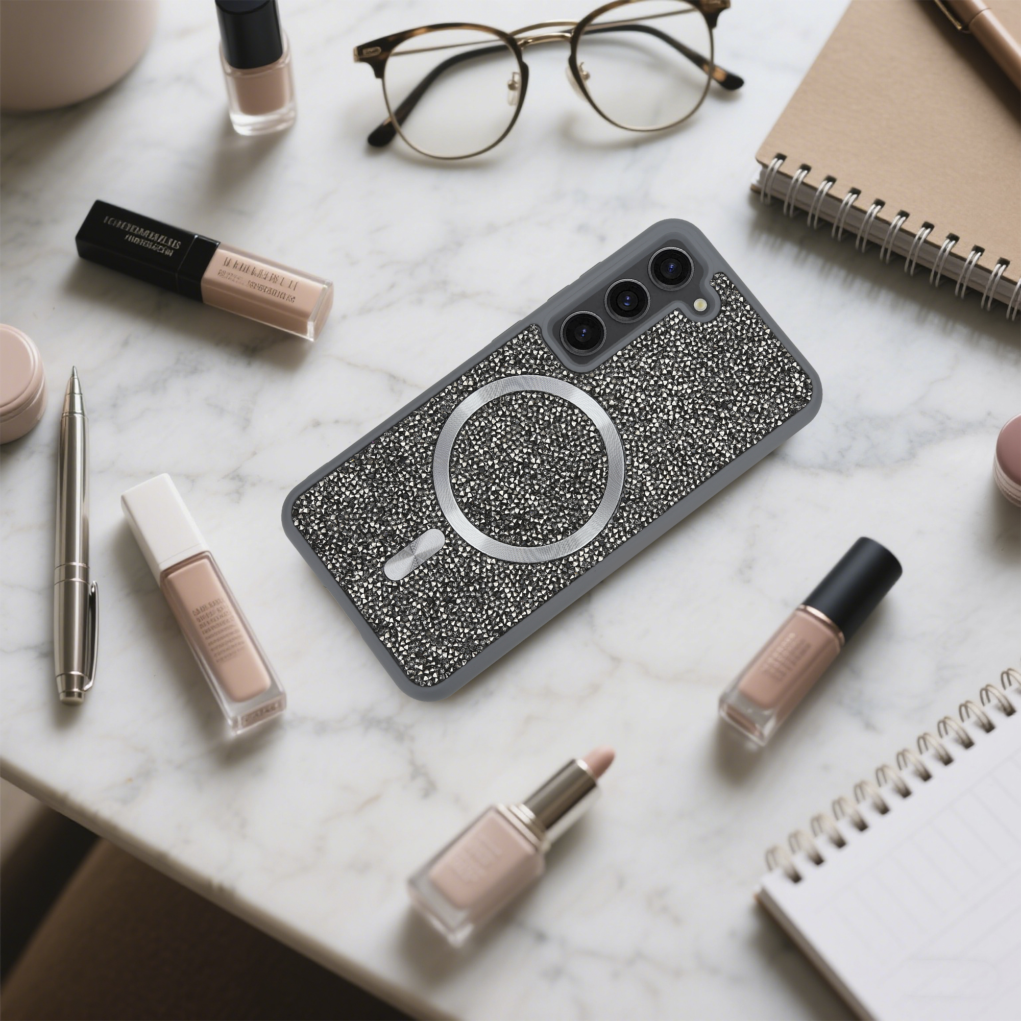 Ein Handycase, Make-up, eine Brille und ein Notizbuch sind auf einer Marmoroberfläche angeordnet. Das Handycase ist mit Kristallen bedeckt.