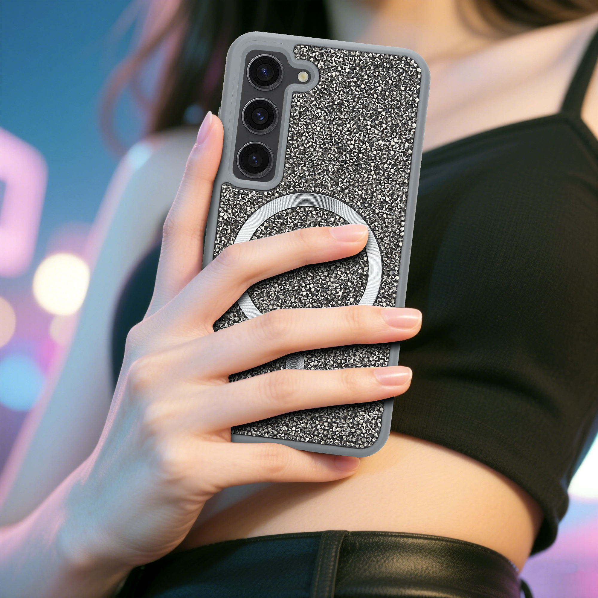 Eine Hand hält ein Telefon mit einer kristallbedeckten Hülle. Das Telefon hat einen silbernen Ring und drei Kameras. Eine Person in einem schwarzen Oberteil.