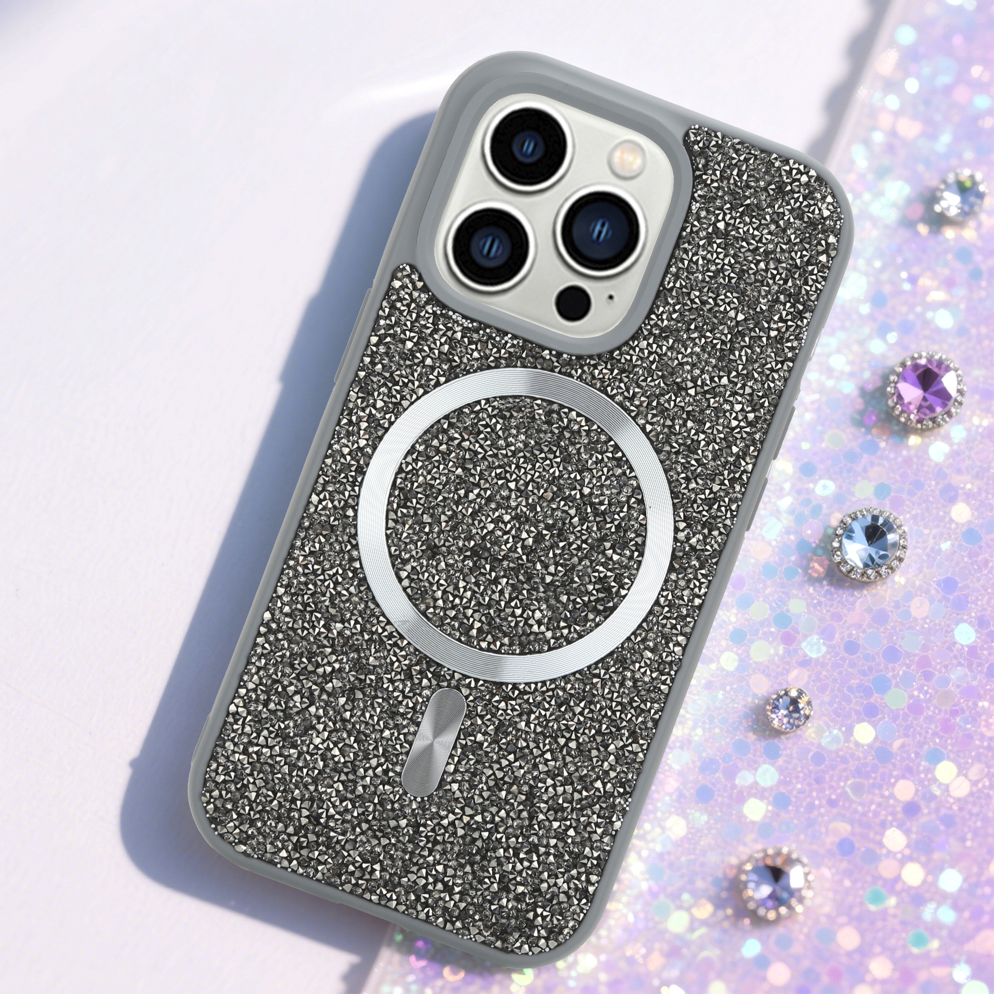 Ein verziertes Handycase mit silbernem Ring, drei Kameralinsen und funkelnden Juwelen auf glitzerndem Hintergrund.