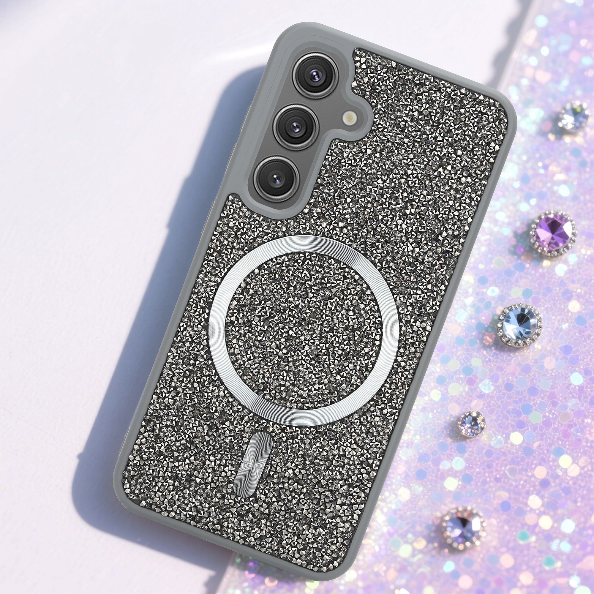 Ein verziertes Handycase mit silbernem Ring und drei Kameralinsen. Glitzernder Hintergrund mit verstreuten Edelsteinen.