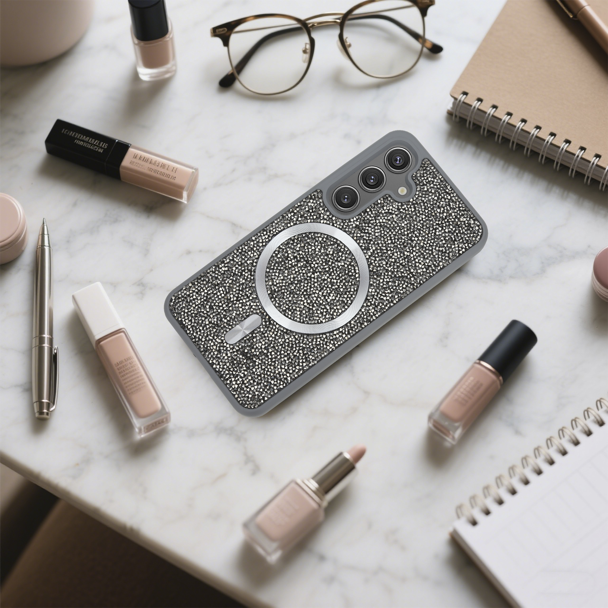 Eine Handyhülle mit Kristallen, Brille, Make-up und ein Notizbuch liegen auf einer Marmoroberfläche.