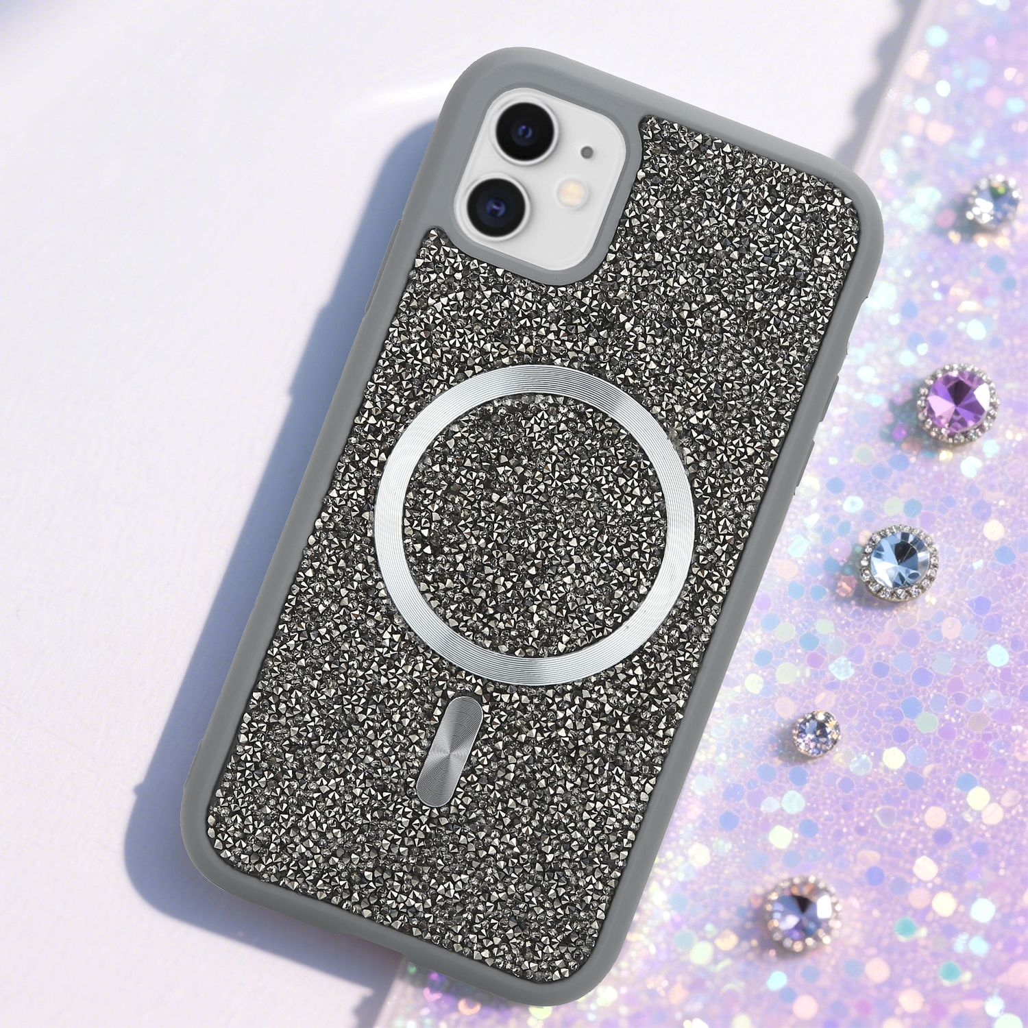 Ein verziertes Handycase mit silbernem Ring und Edelsteinen auf glitzernder Oberfläche. Das Case ist grau mit schwarzer Kamera.