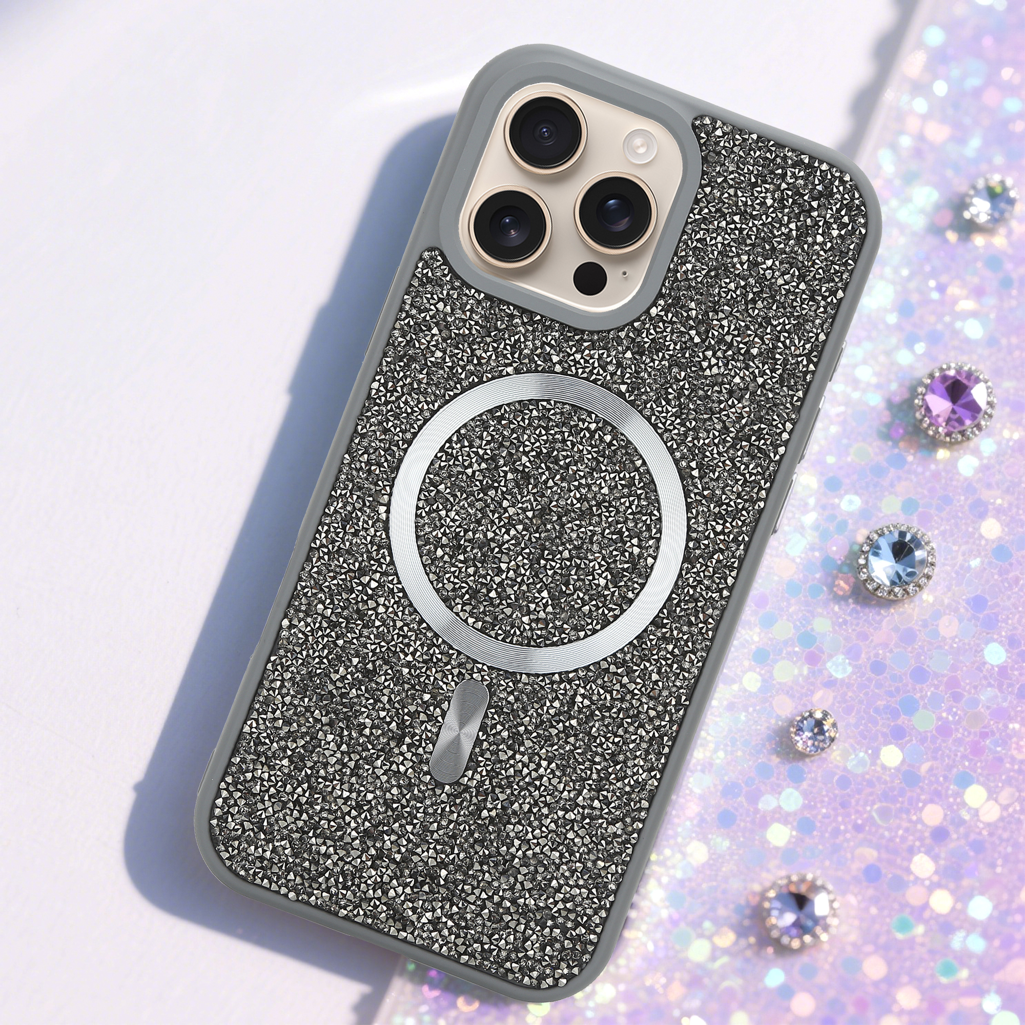 Silbernes, mit Strasssteinen besetztes Handycase mit MagSafe-Ring. Strasssteine auf pinkfarbenem Glitzerhintergrund mit Juwelen.