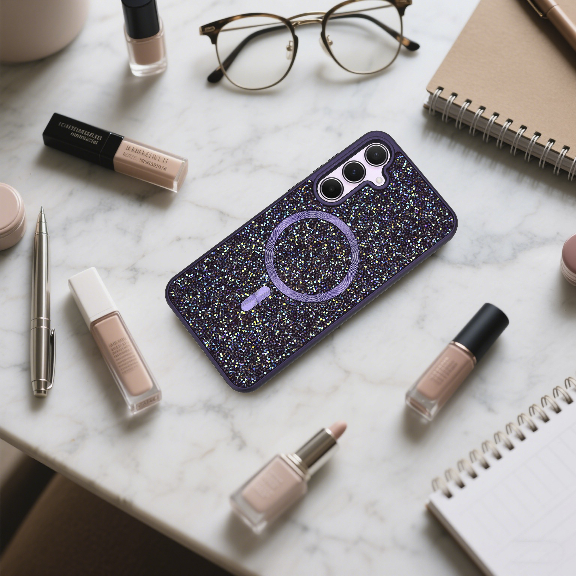 Eine Handyhülle mit Kristallen, Brille, Make-up und ein Notizbuch liegen auf einer Marmoroberfläche.