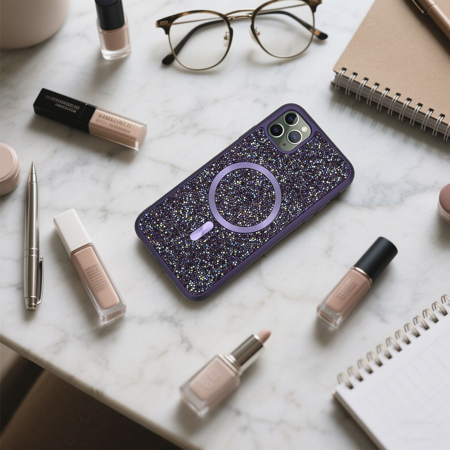 Ein glitzerndes Handycase ist von Make-up, Brille, Notizbuch und Stift auf einer Marmoroberfläche umgeben.