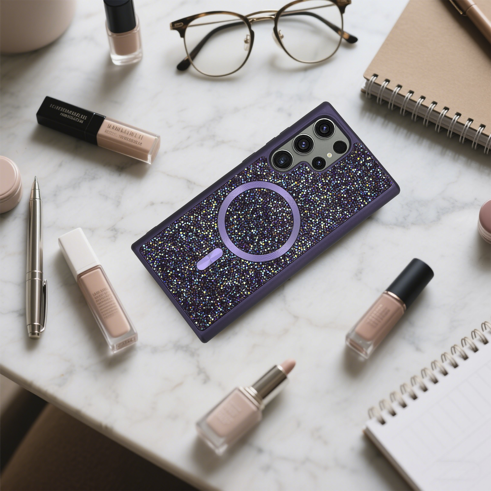 Lila Handyhülle mit Kristallen, umgeben von Make-up, Brille, Stift und Notizbuch auf Marmor.