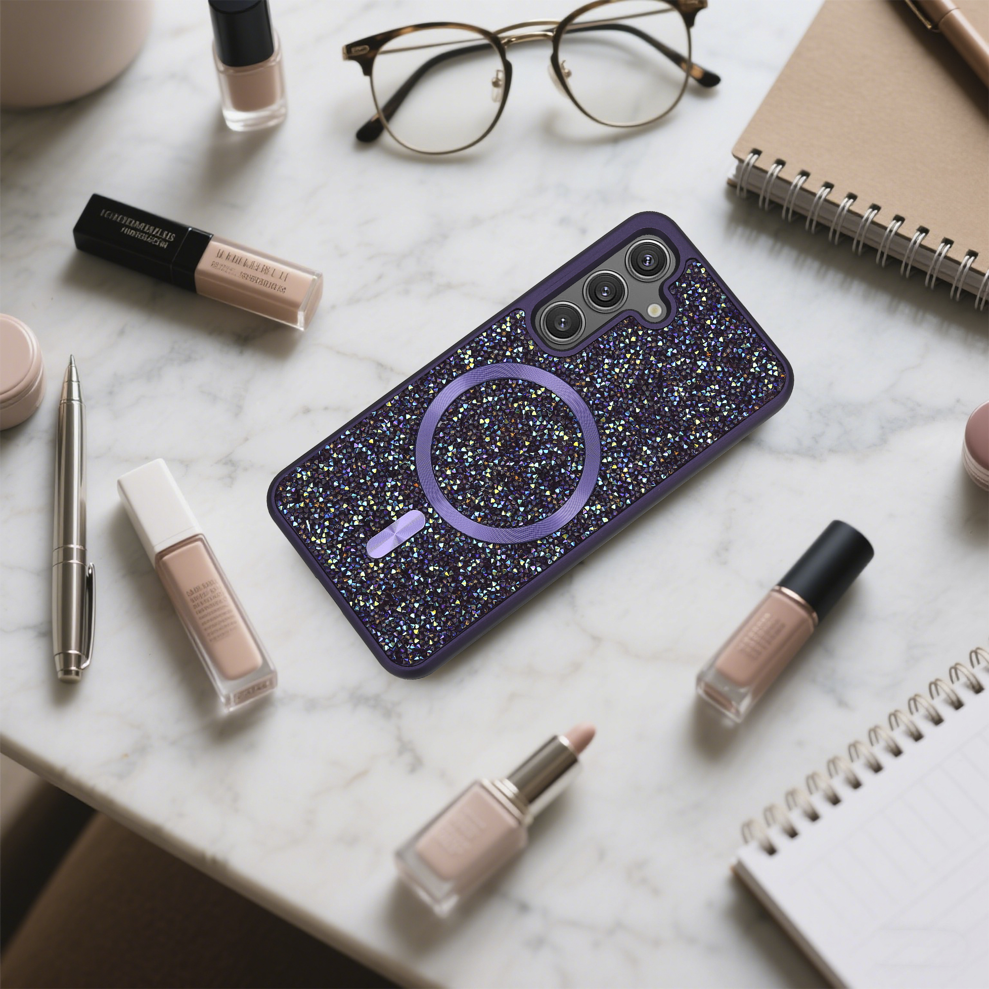 Ein verziertes Handycase, Brille, Kosmetika, Stift und Notizbuch liegen auf Marmor. Eine elegante Komposition.