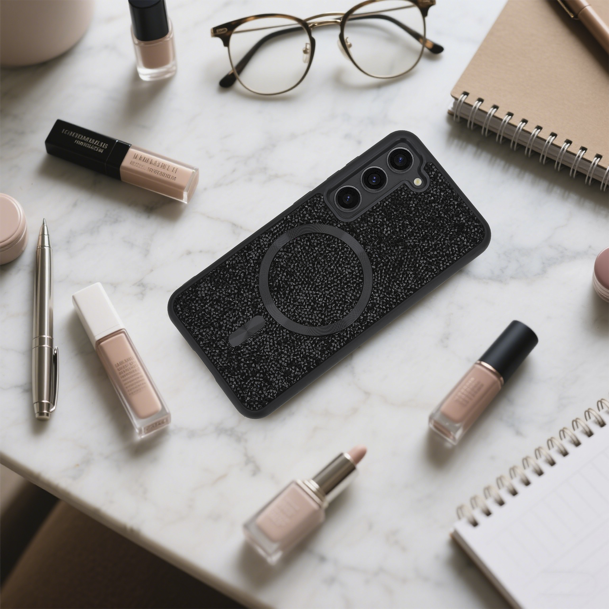 Eine Handyhülle, Make-up, eine Brille und ein Notizbuch liegen auf einer Marmoroberfläche.