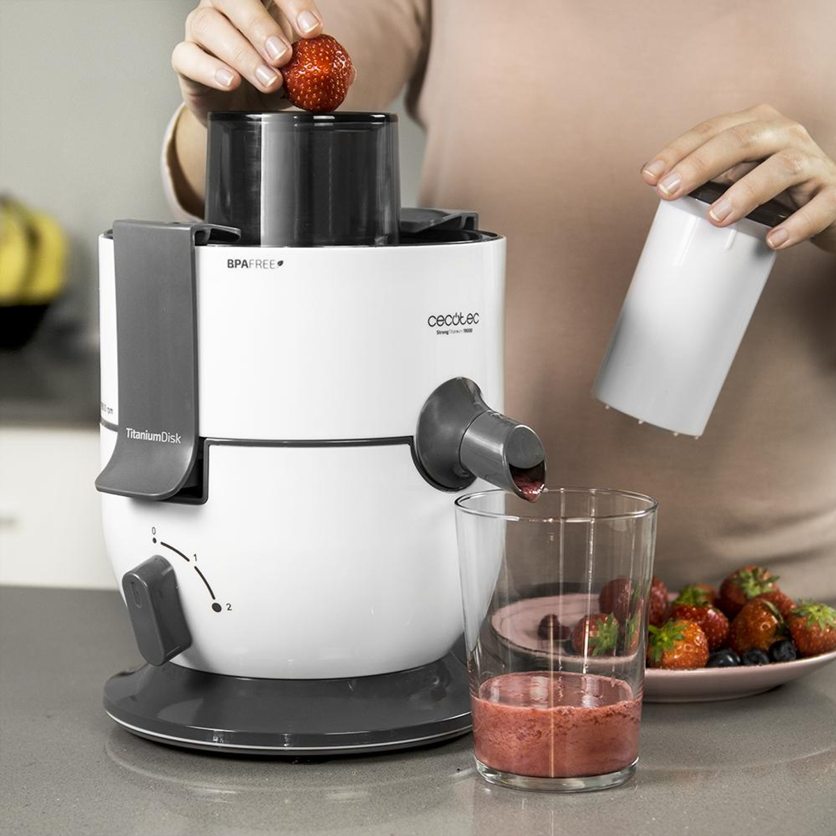 Una persona sta facendo succo di fragole con un estrattore bianco e grigio. Un bicchiere con succo, fragole e mirtilli sono visibili.