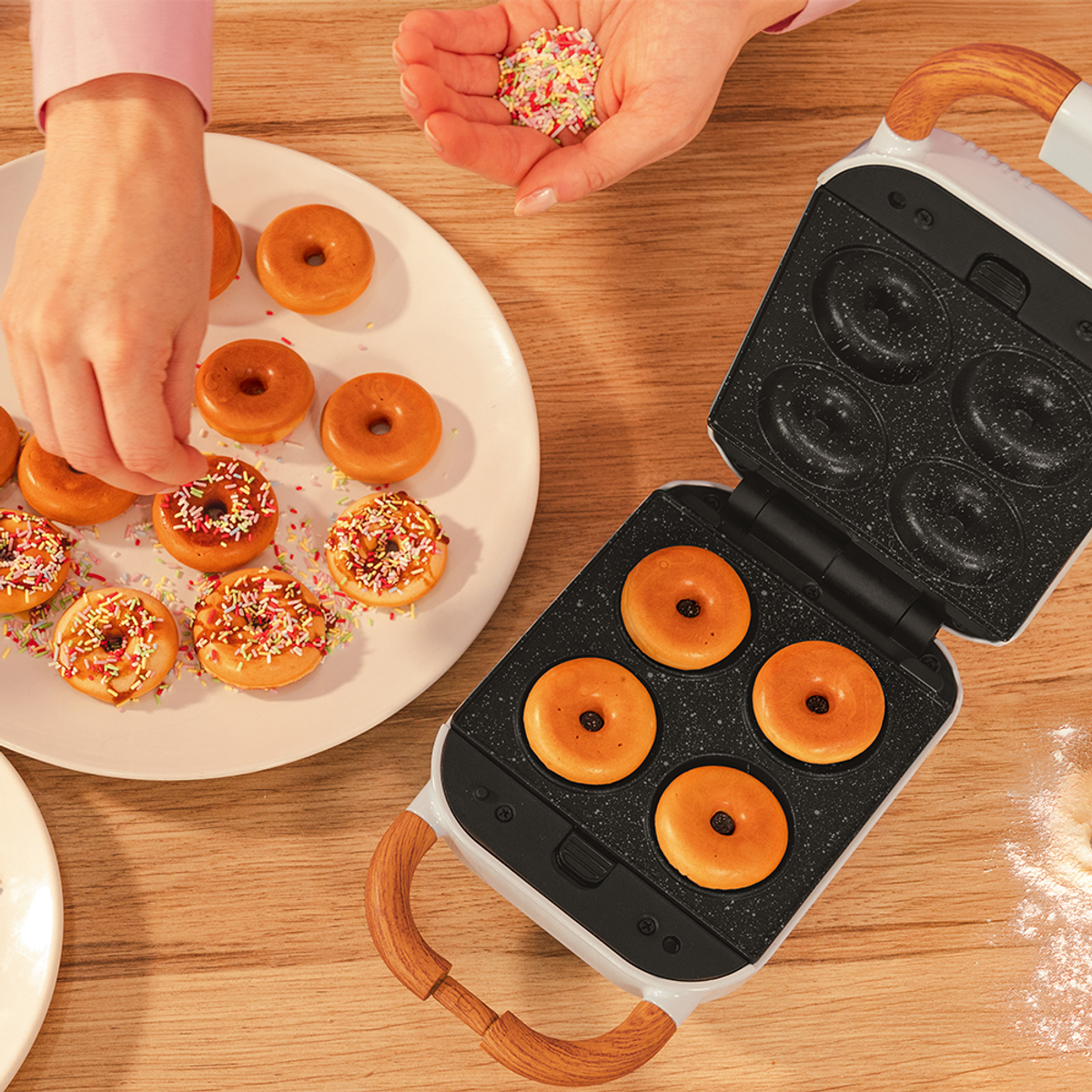 Manos decorando donas con chispas. Algunas donas están en un plato, otras en una máquina para hacer donas. Mesa de madera al fondo.