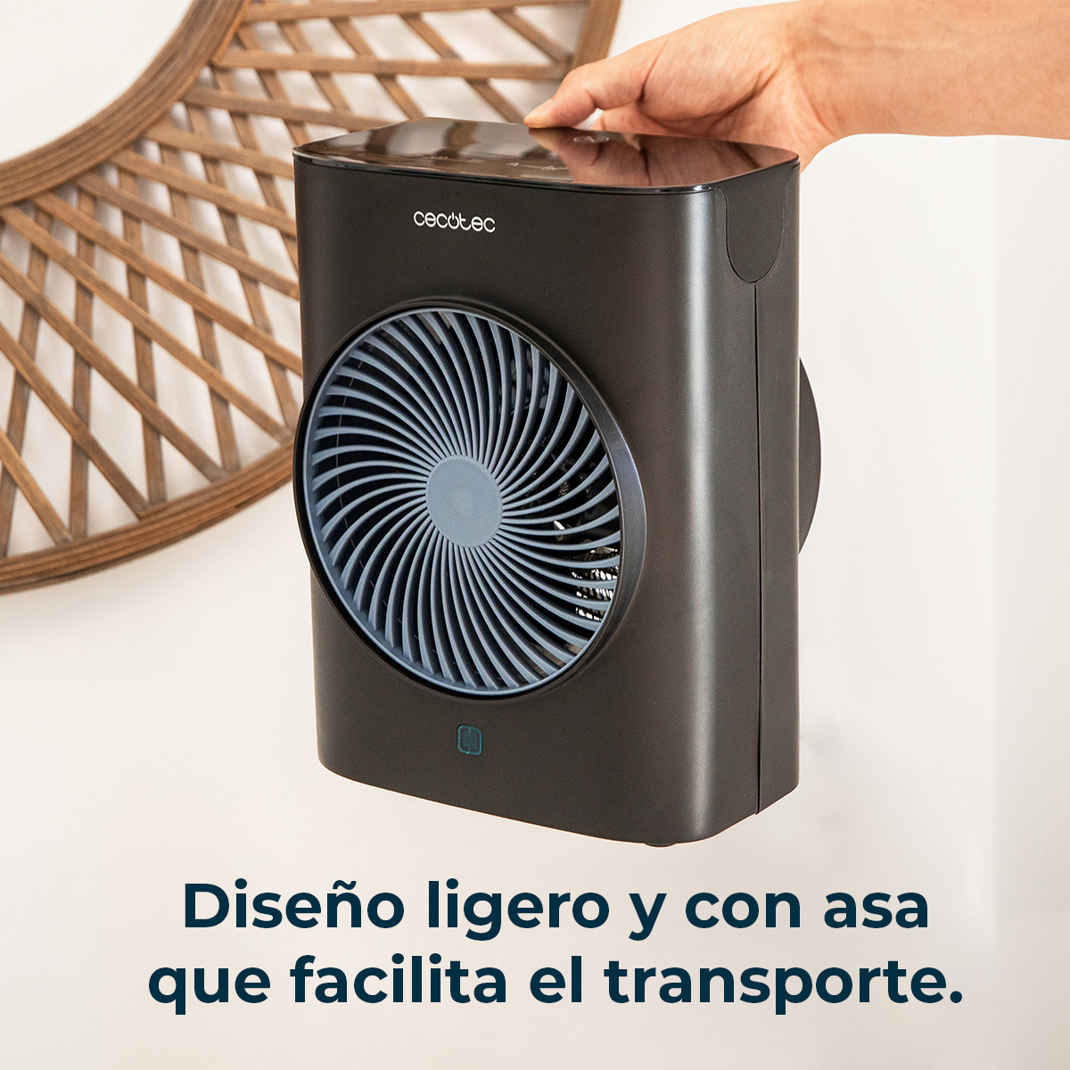 Un ventilatore nero è tenuto in mano. Ha una pala grigia. Lo sfondo ha un pezzo di legno decorativo e un muro bianco.