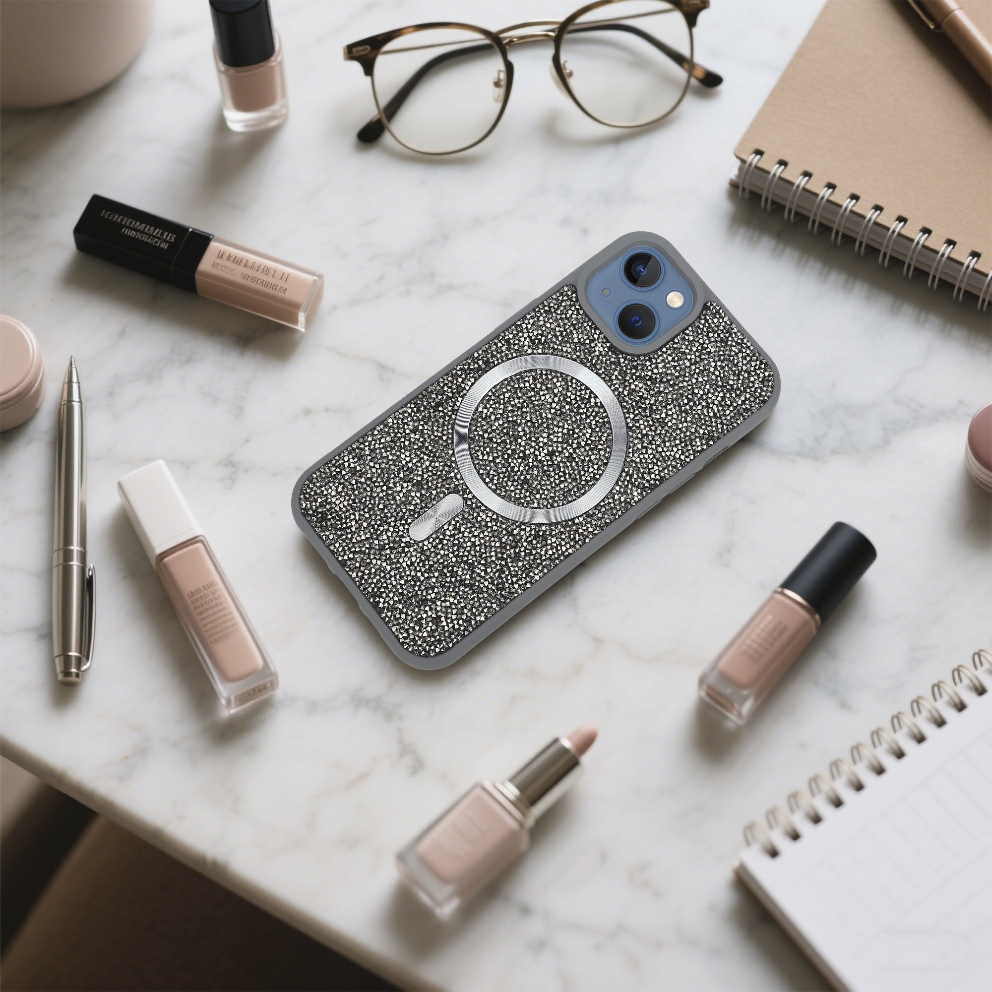 Ein iPhone mit Strass-Hülle, Make-up, Brille und Notizbuch liegen auf Marmor. Lippenstift im Vordergrund.