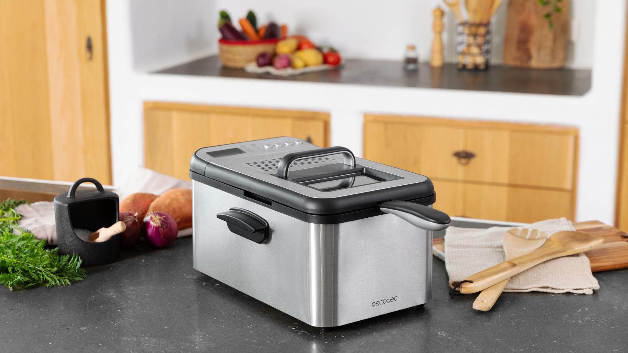 Frytownica ze stali nierdzewnej na blacie. Obecne są również warzywa i drewniane przybory. Tło obejmuje szafki kuchenne.