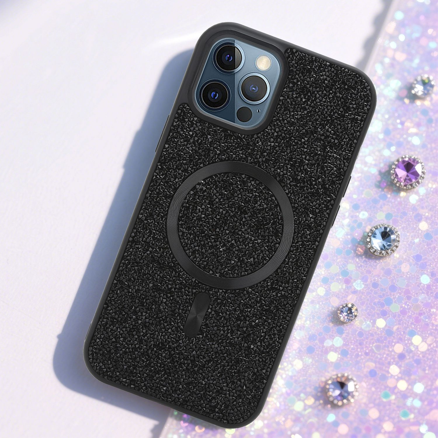 Schwarze, strukturierte Handyhülle mit kreisförmigem Magnetring und Kameralinsen. Glitzernder Hintergrund mit verstreuten Juwelen.