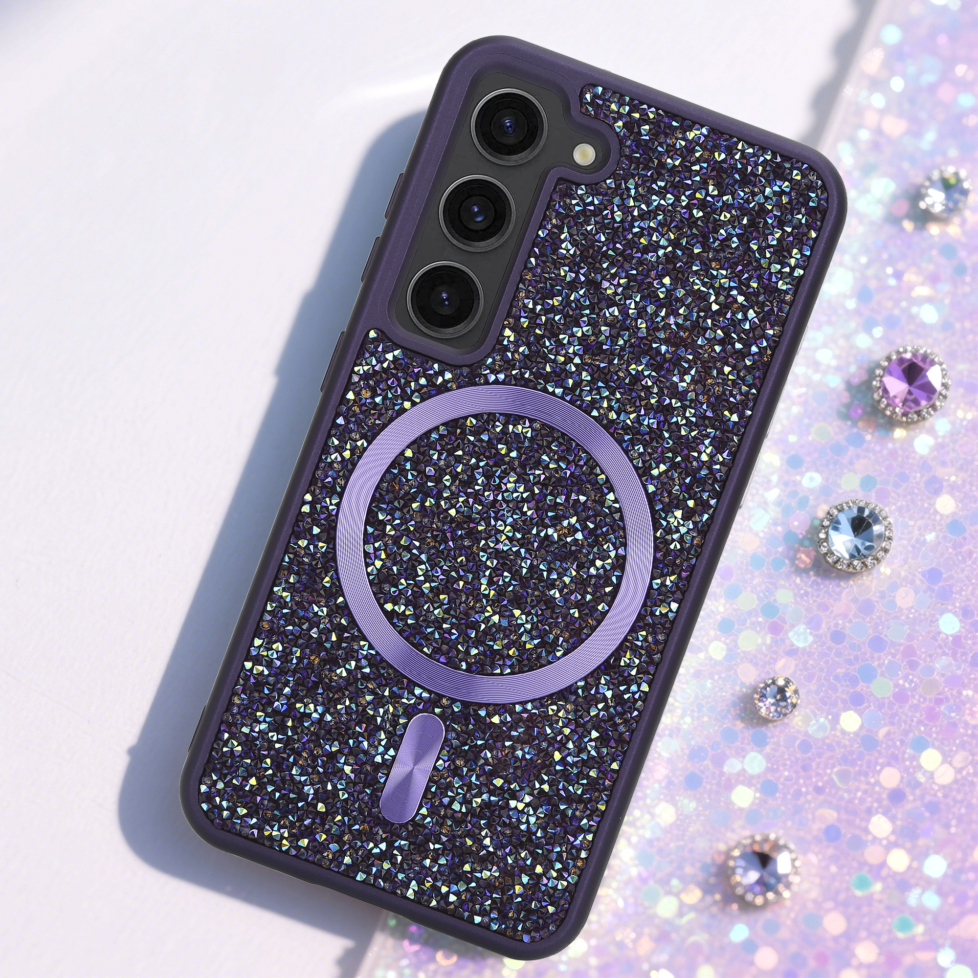 Eine mit Glitzersteinen besetzte Handyhülle mit lilafarbenen Akzenten und einem Magnetring. Mehrere Edelsteine ​​liegen auf einer glitzernden Oberfläche.