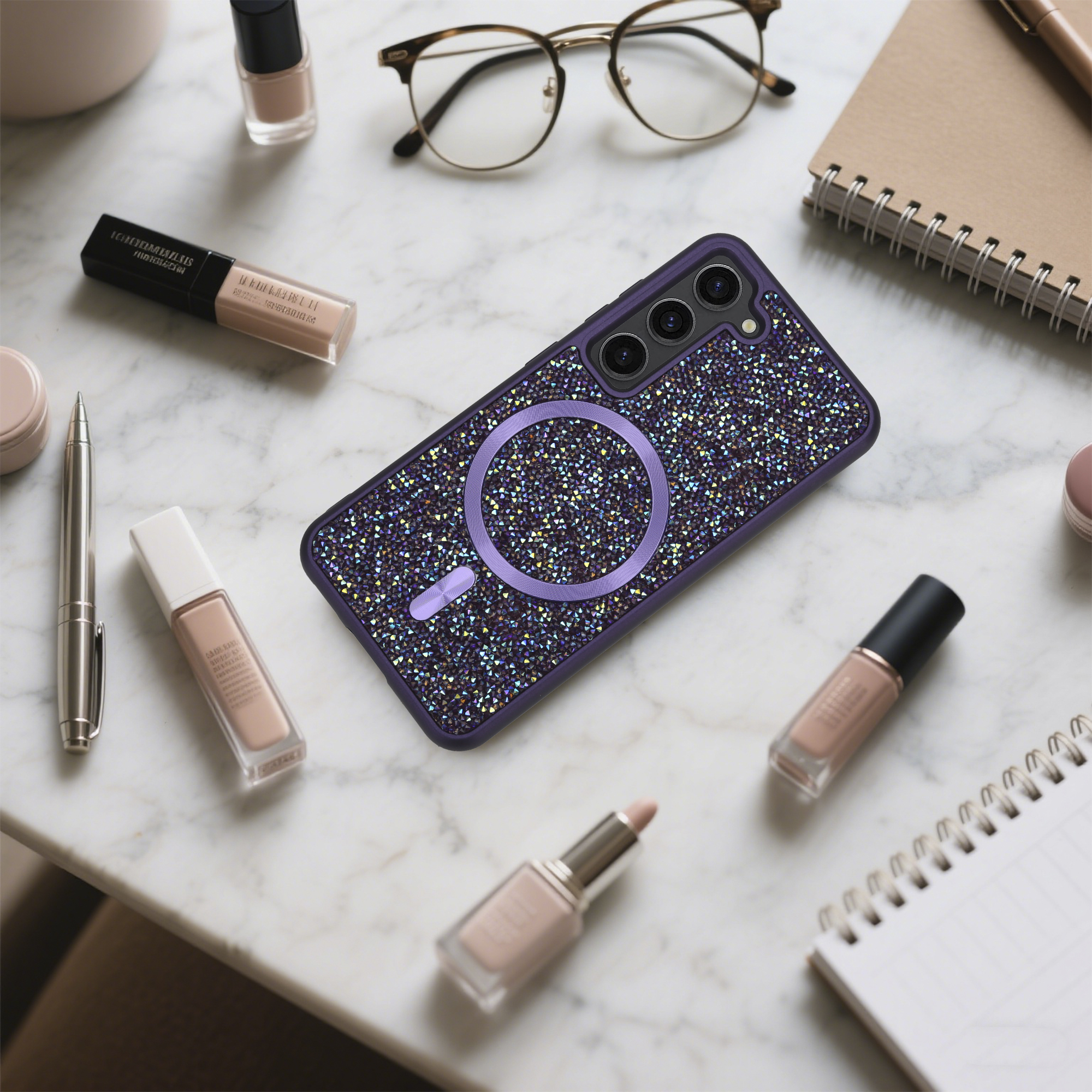 Eine Handyhülle mit Kristallen, Make-up, Brille und Notizbuch liegen auf einer Marmoroberfläche, verschwommener Hintergrund.