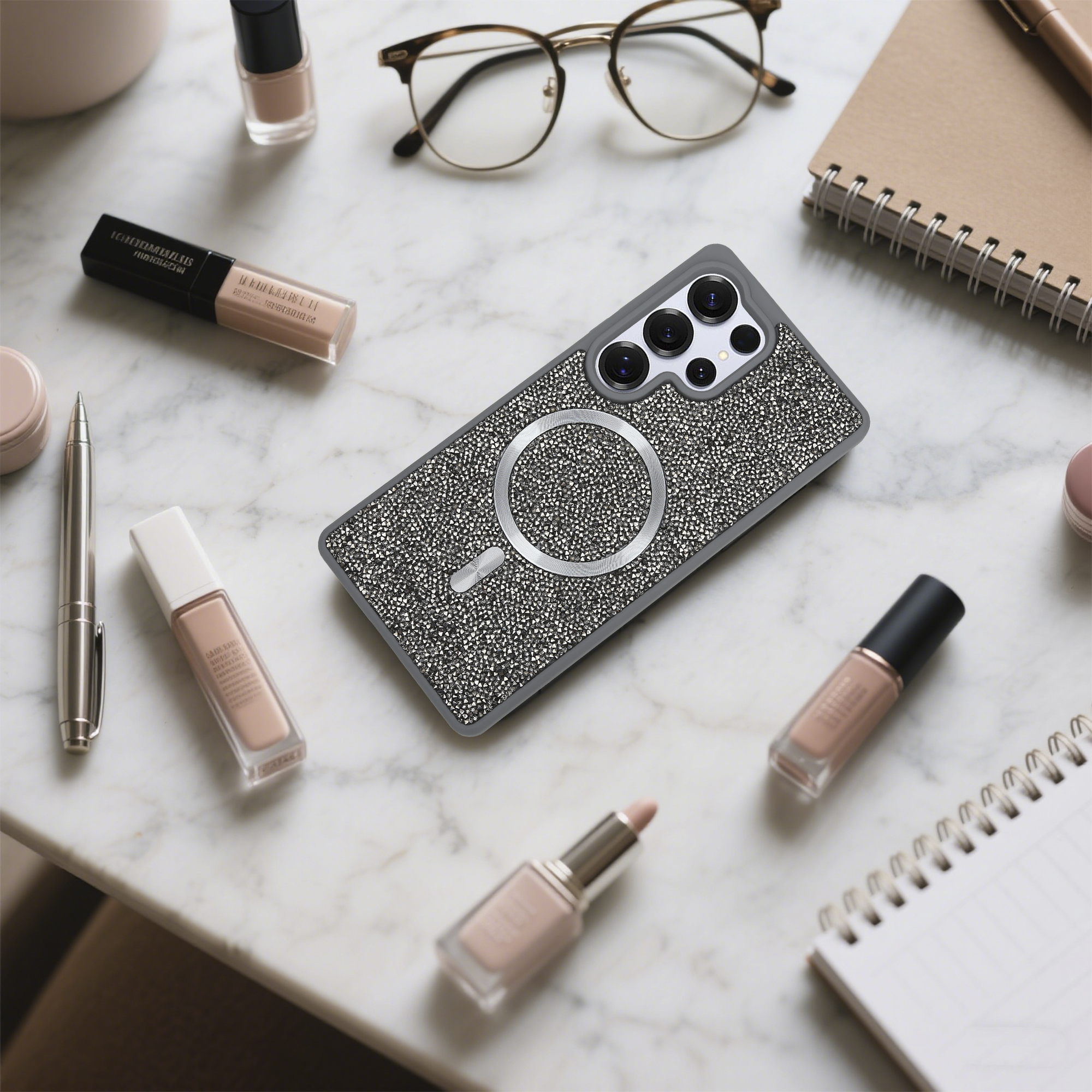 Eine Handyhülle mit Kristallen, Make-up, Brille, Stift und Notizblock liegen auf einer Marmoroberfläche.