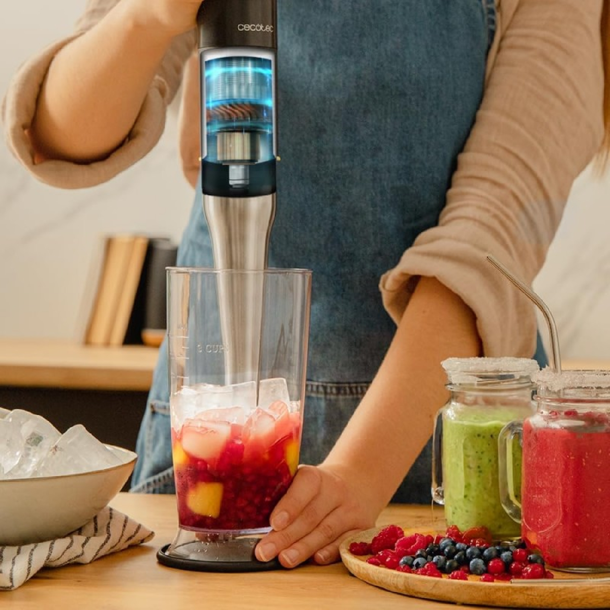 Osoba miksuje napój blenderem ręcznym. Szklanka zawiera lód i owoce. Inne zmiksowane napoje i owoce na stole.