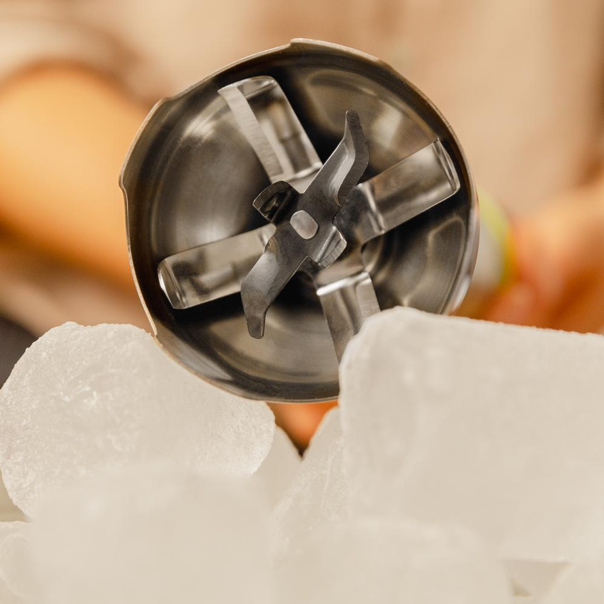 Zbliżenie głowicy blendera z ostrzami, spoczywającej na kostkach lodu. Ostrza znajdują się na środku, otoczone lodem.