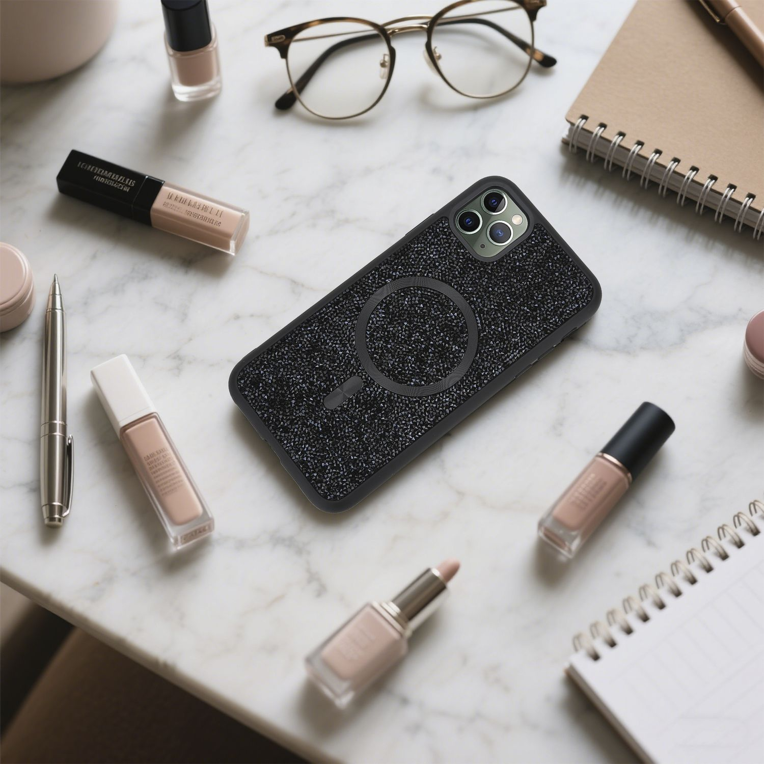 Ein Handycase, Brille, Make-up, Stift und Notizbuch liegen auf einer Marmoroberfläche. Das Handycase ist schwarz mit glitzernder Textur.
