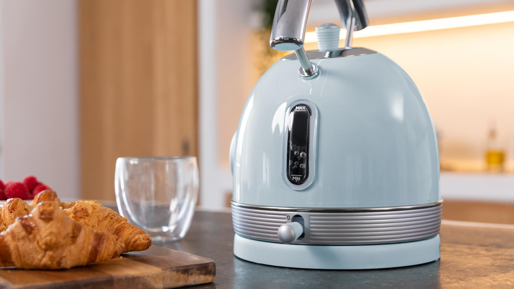 Jasnoniebieski czajnik, rogaliki, maliny i szklanka na blacie. Tło przedstawia drewniane drzwi i butelkę.