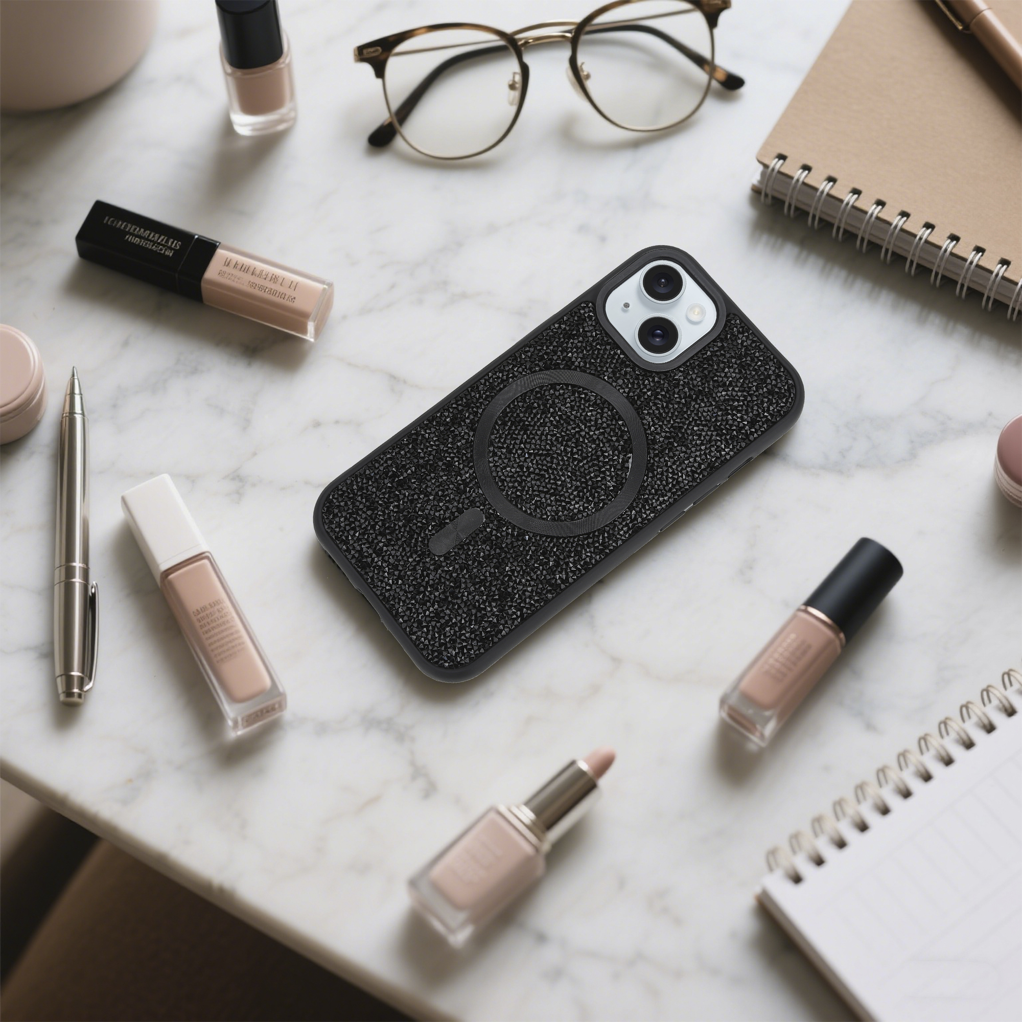 Ein Telefon in einer schwarzen, glitzernden Hülle liegt auf einer Marmoroberfläche, umgeben von Make-up, Brille und einem Notizbuch.