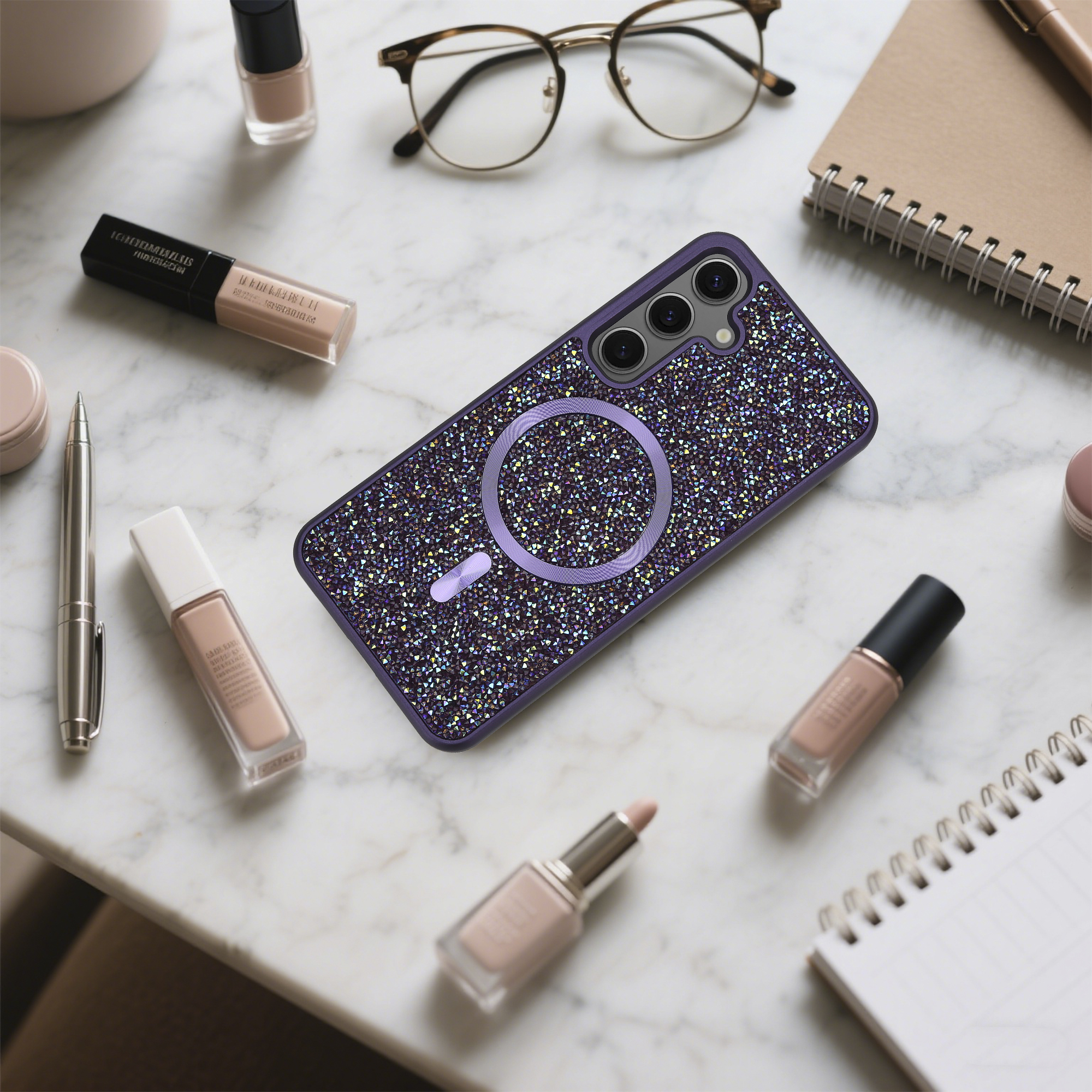 Ein mit Kristallen bedecktes Telefoncase, umgeben von Make-up, Brille, Notizbuch und Stift auf Marmor.