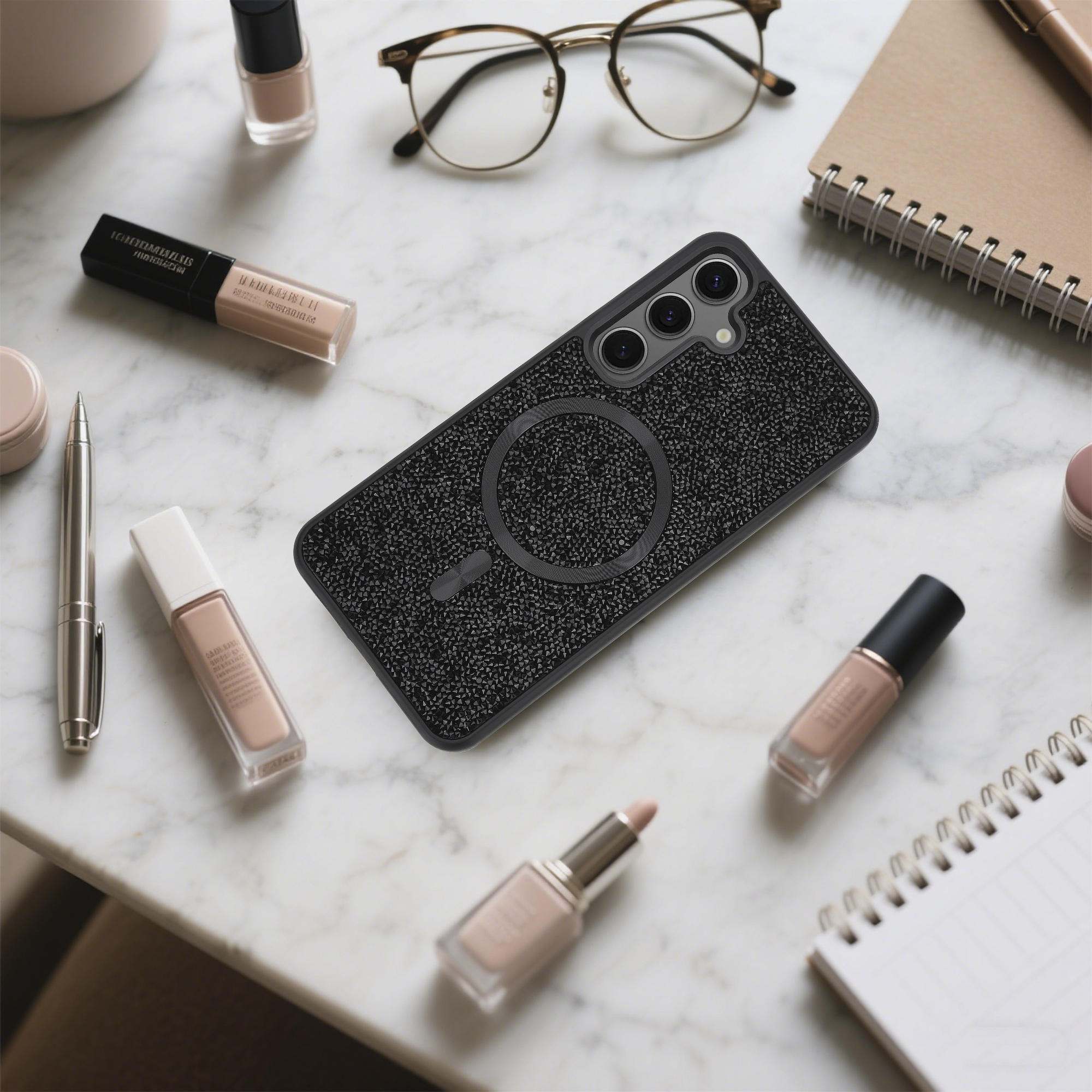 Auf einer Marmoroberfläche sind eine Handyhülle, Make-up, eine Brille, ein Stift und ein Notizbuch angeordnet. Die Handyhülle ist schwarz mit Glitzer.