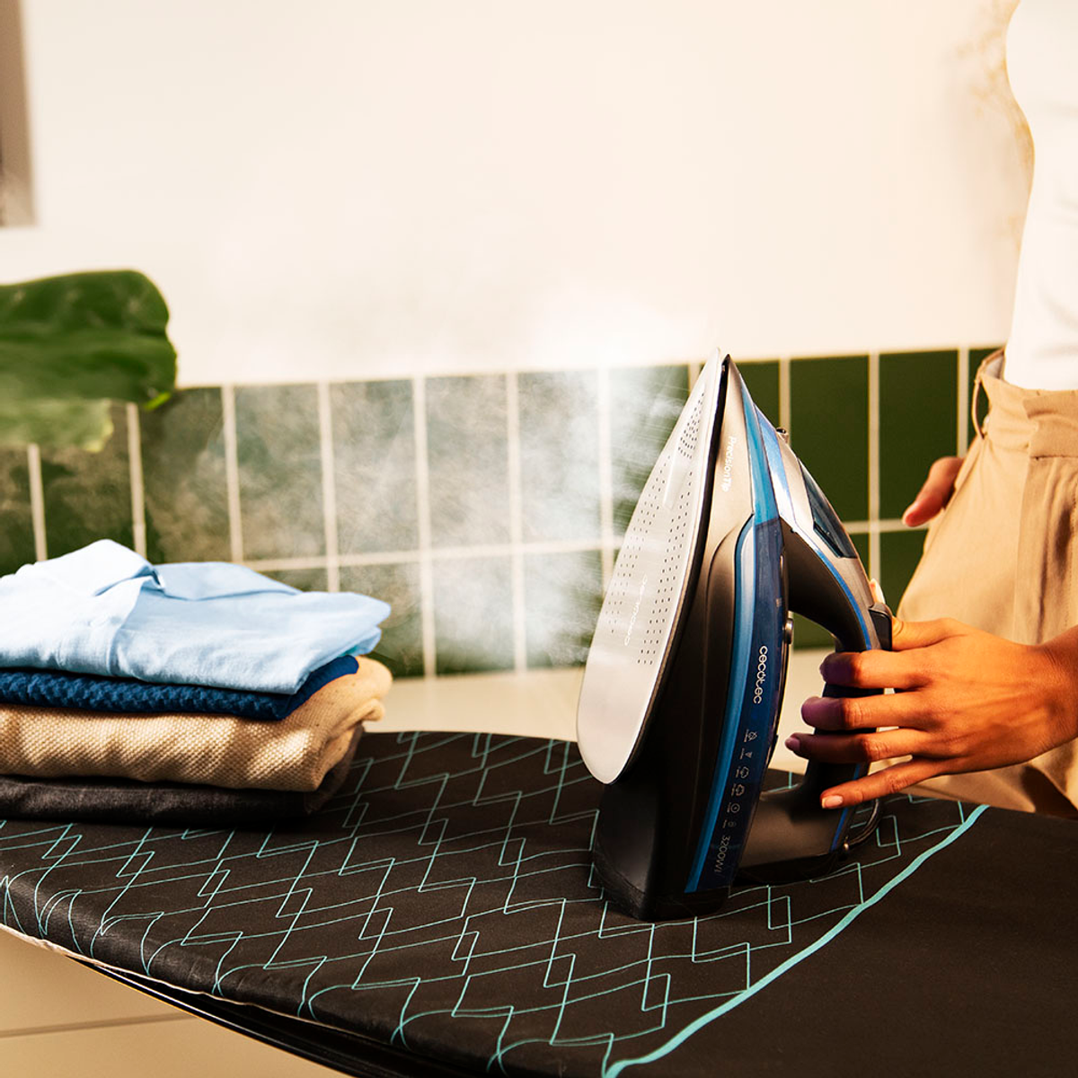 Osoba prasuje ubrania żelazkiem parowym na desce do prasowania. Na desce leży stos złożonych koszul.