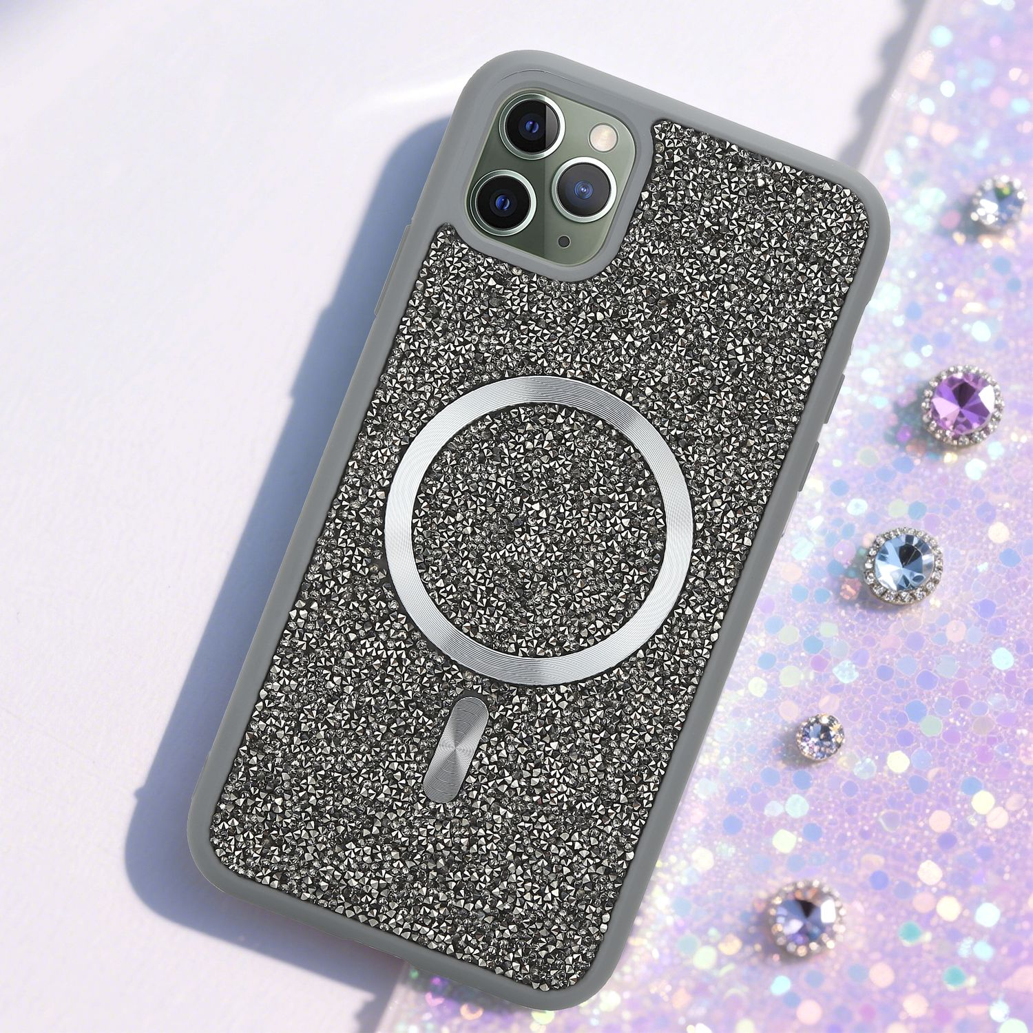 Ein mit Strass besetztes Handycase mit einem silbernen Ring und Edelsteinen. Der Hintergrund ist eine glitzernde, hellviolette Oberfläche mit verstreuten Juwelen.