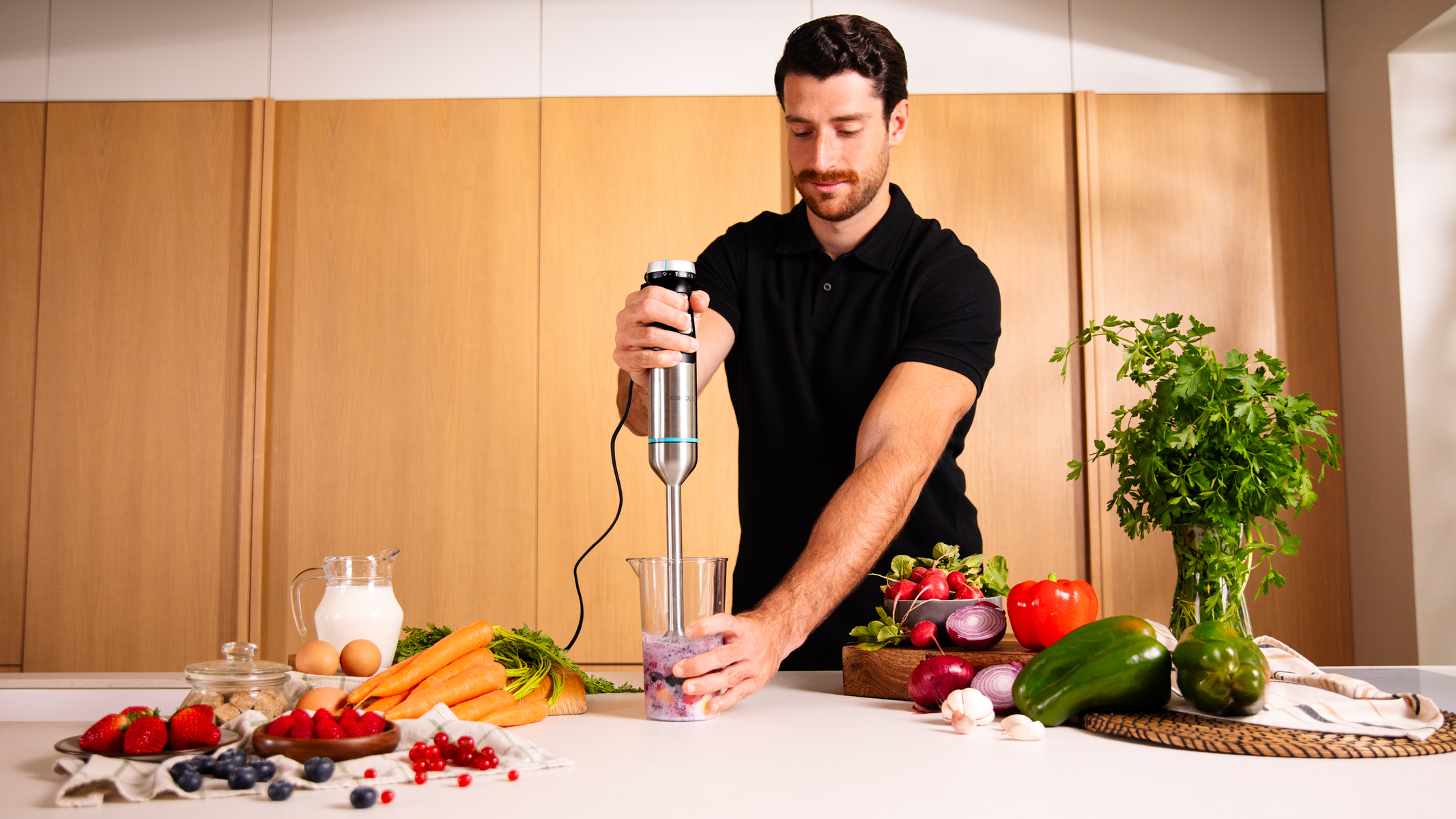 Un uomo frulla frutta in cucina. Ingredienti come bacche, verdure e latte sono sul bancone. Mobili in legno sullo sfondo.