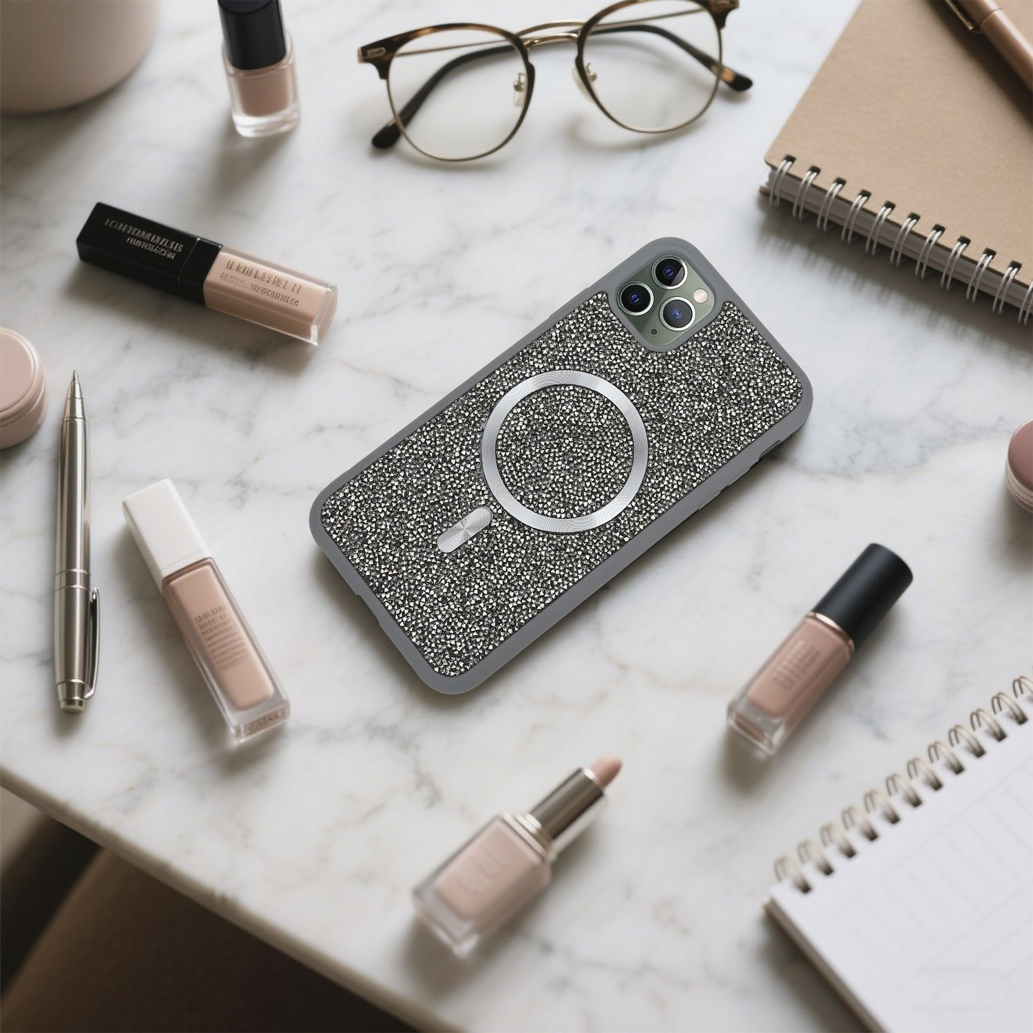 Ein Telefon mit Strasssteinen ist von Make-up, Brille, Stift und Notizblock auf einer Marmoroberfläche umgeben.