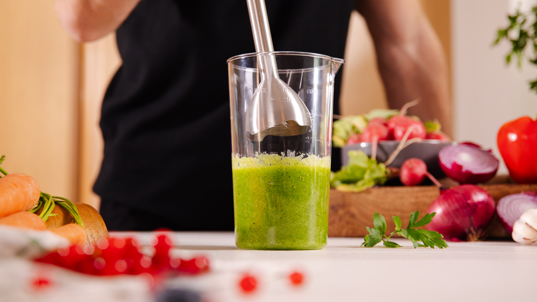 Un frullatore in un bicchiere con liquido verde. Sullo sfondo ci sono verdure come carote, cipolle e ravanelli.