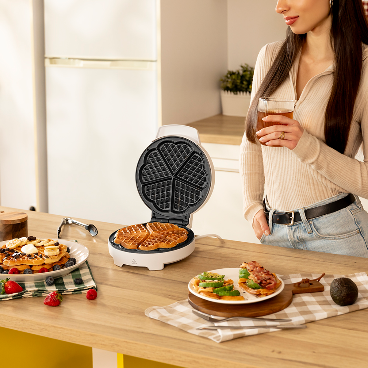 Una donna con un bicchiere di tè è in piedi vicino a un tavolo con waffle, una macchina per waffle e piatti di cibo. Sono visibili bacche e avocado.