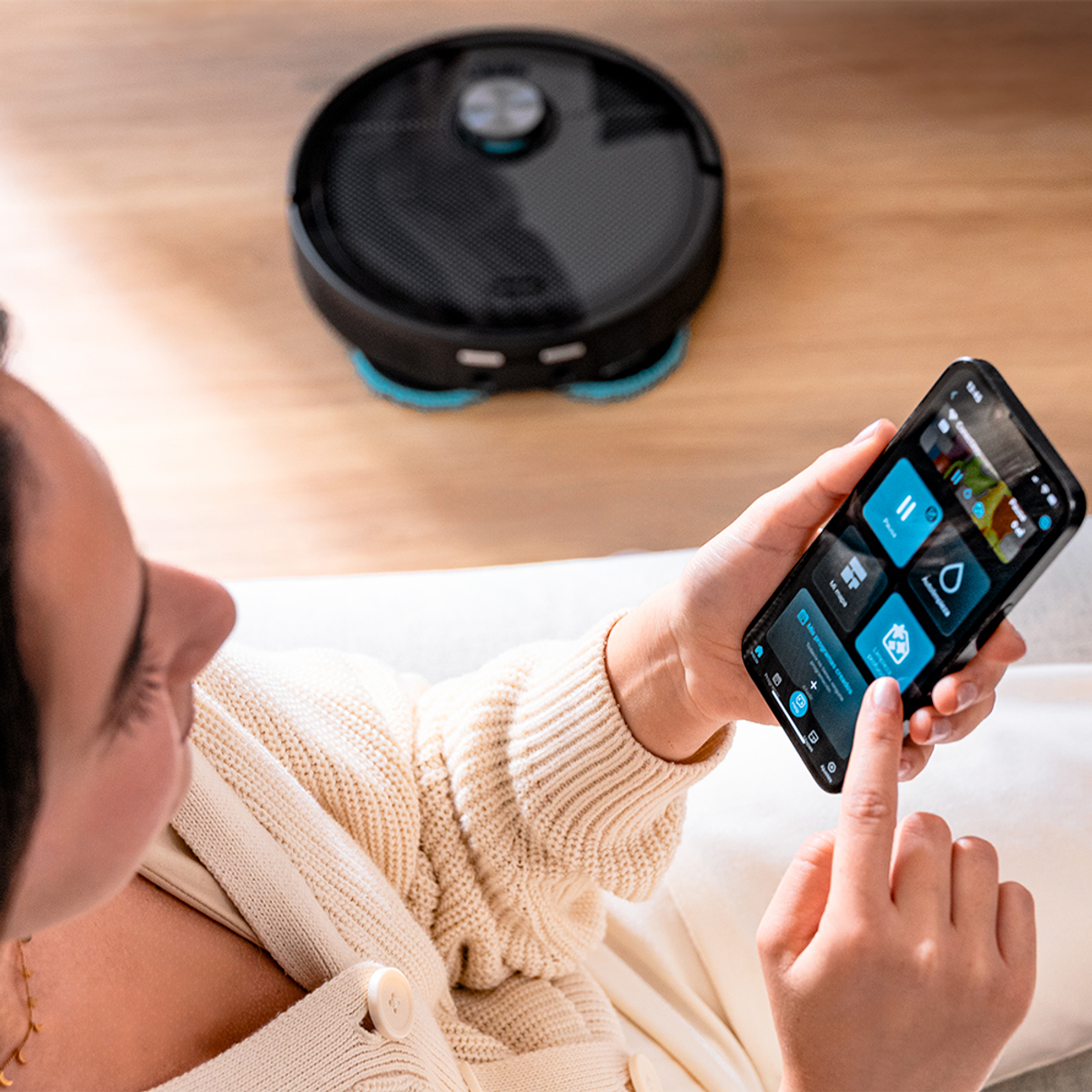 Una persona usa un teléfono para controlar una aspiradora robot negra. El teléfono muestra controles de la aplicación. Se ve un suéter blanco.