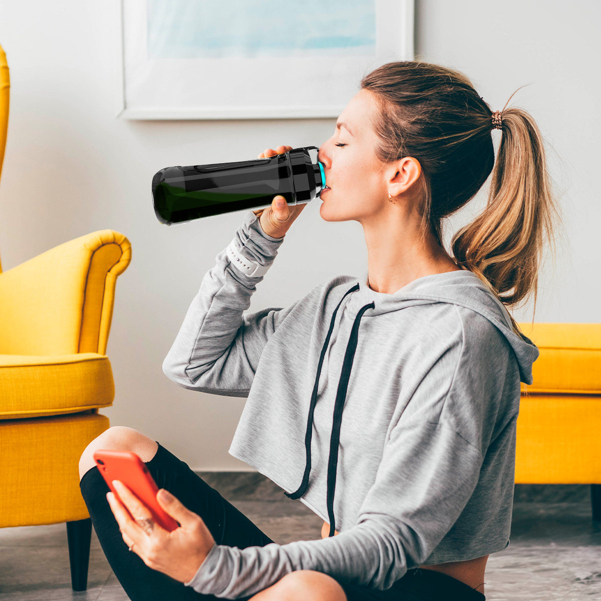 Vrouw drinkt uit een zwarte fles, met een rode telefoon. Gele stoel en poef op de achtergrond.